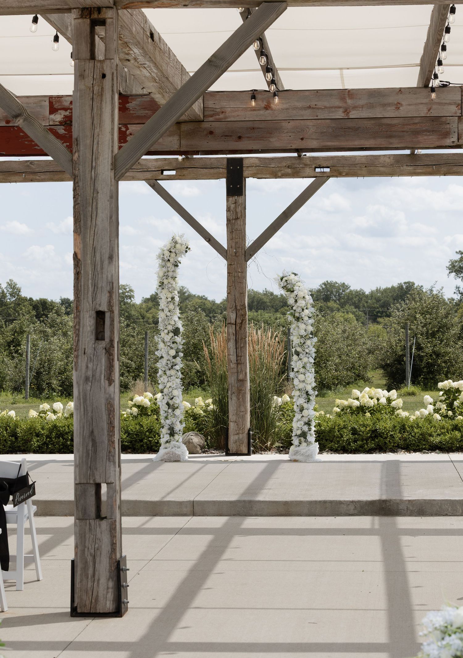 Rustic wooden pergola with white floral columns framing an outdoor wedding ceremony space overlooking lush greenery.
