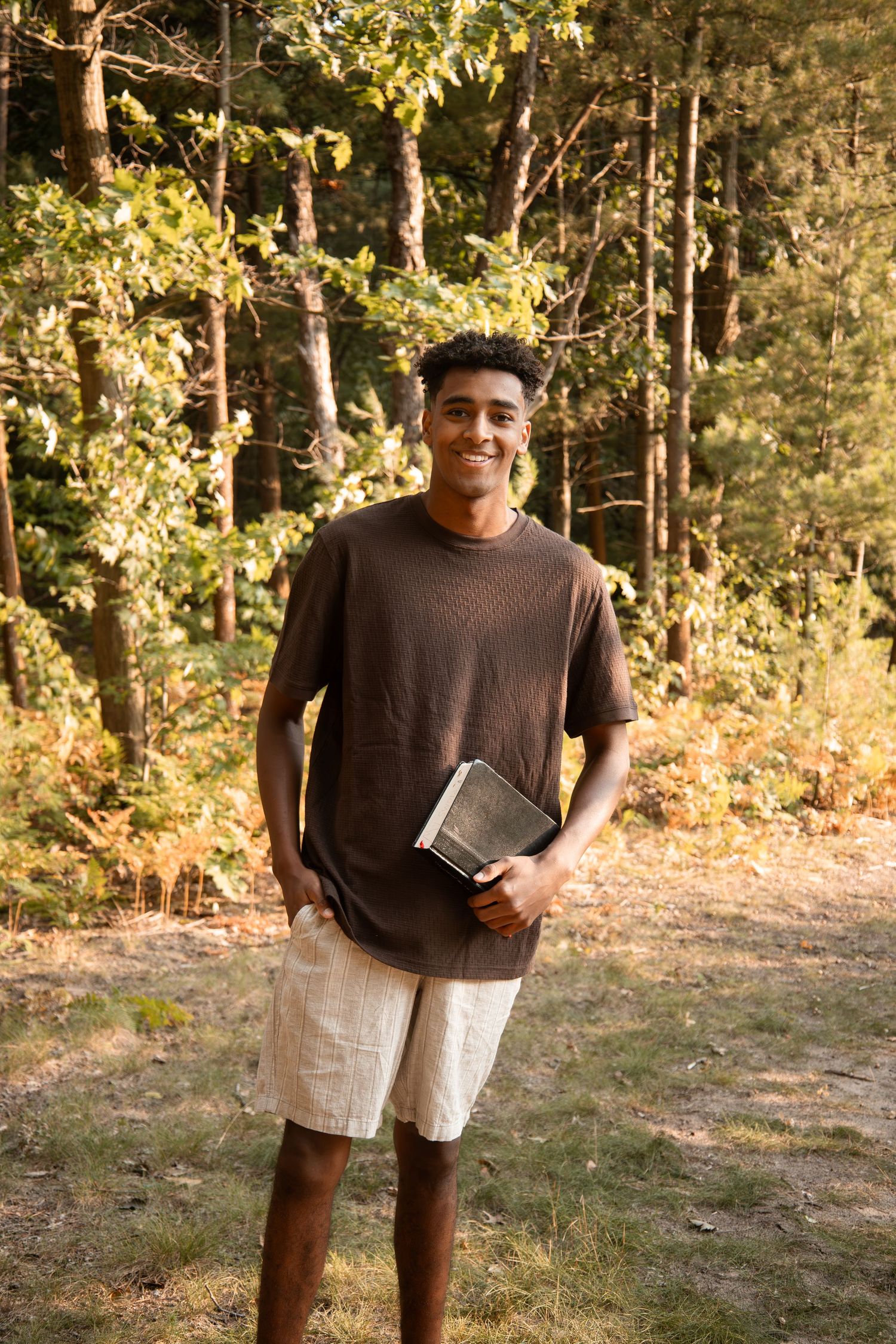 Young Black man smiling outdoors, holding a book, wearing brown t-shirt and khaki shorts with forest background.