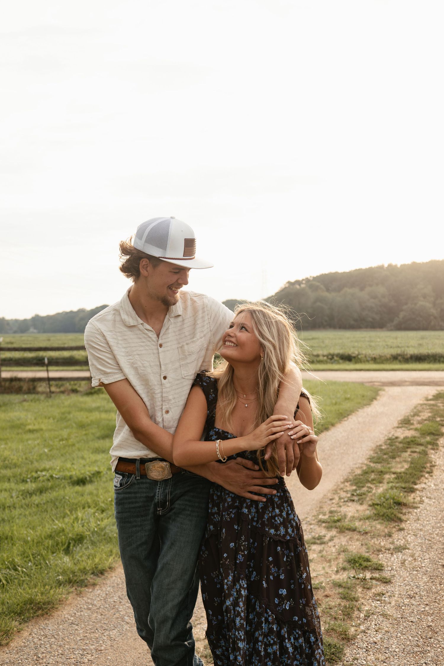 A couple embraces on a rural dirt path surrounded by green fields at golden hour, smiling at each other.