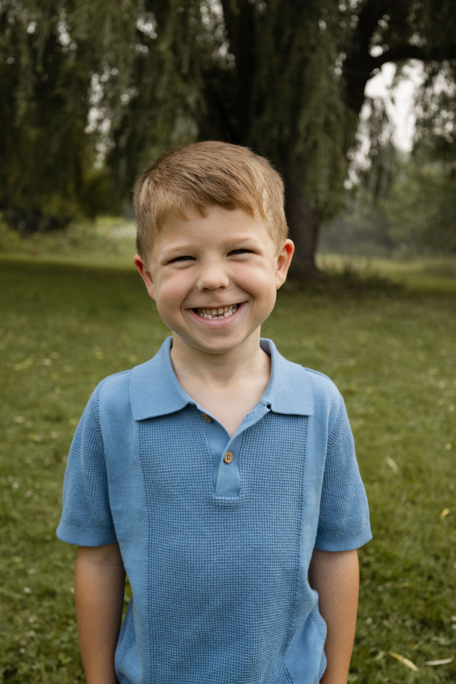 A smiling young boy with red hair wearing a blue polo shirt standing outdoors in a green park.