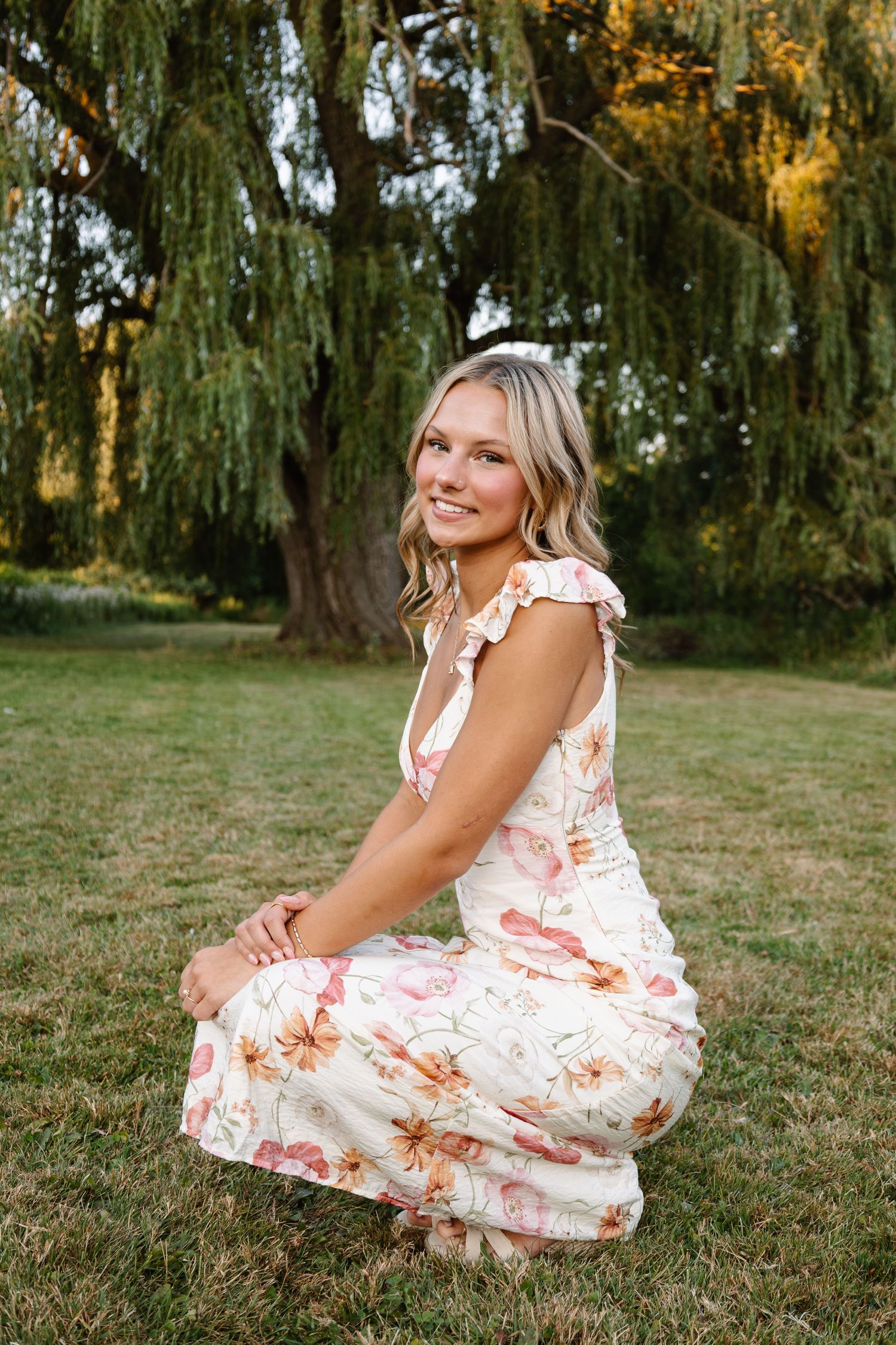 Young woman in floral dress sitting on grass, smiling outdoors near a weeping willow tree.