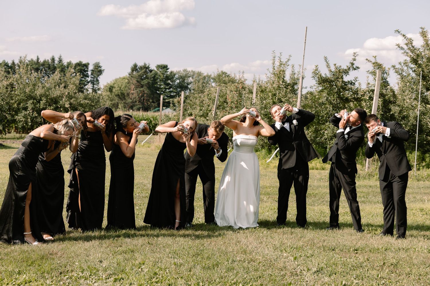 Wedding party in black attire flanking bride and groom outdoors, all covering their faces playfully in a green field.