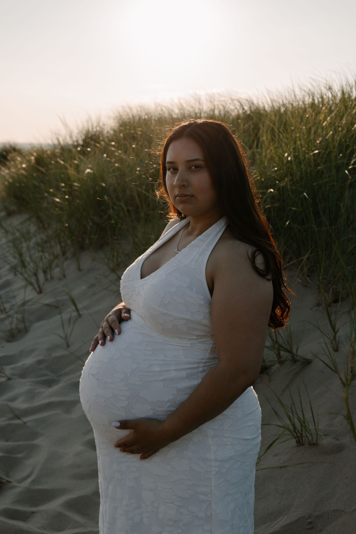 Pregnant woman in white dress holding belly on sandy beach dunes at golden hour sunset.