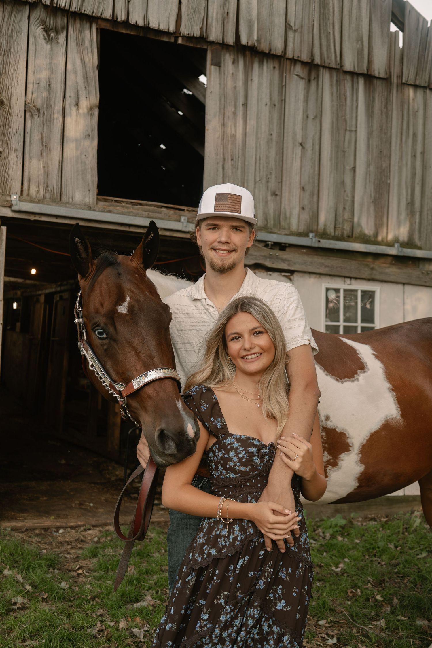 A smiling couple poses with a brown and white paint horse in front of a rustic wooden barn.