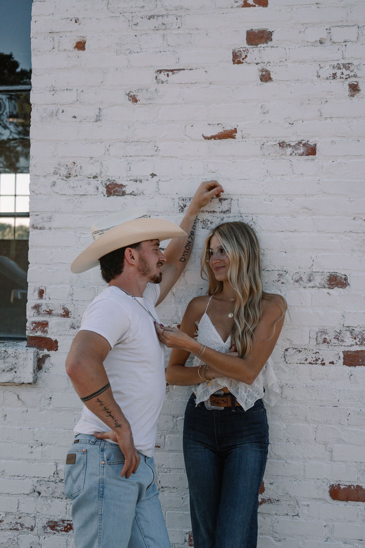 A couple in Western style outfits poses playfully against a white brick wall, man in cowboy hat and woman in lace top.