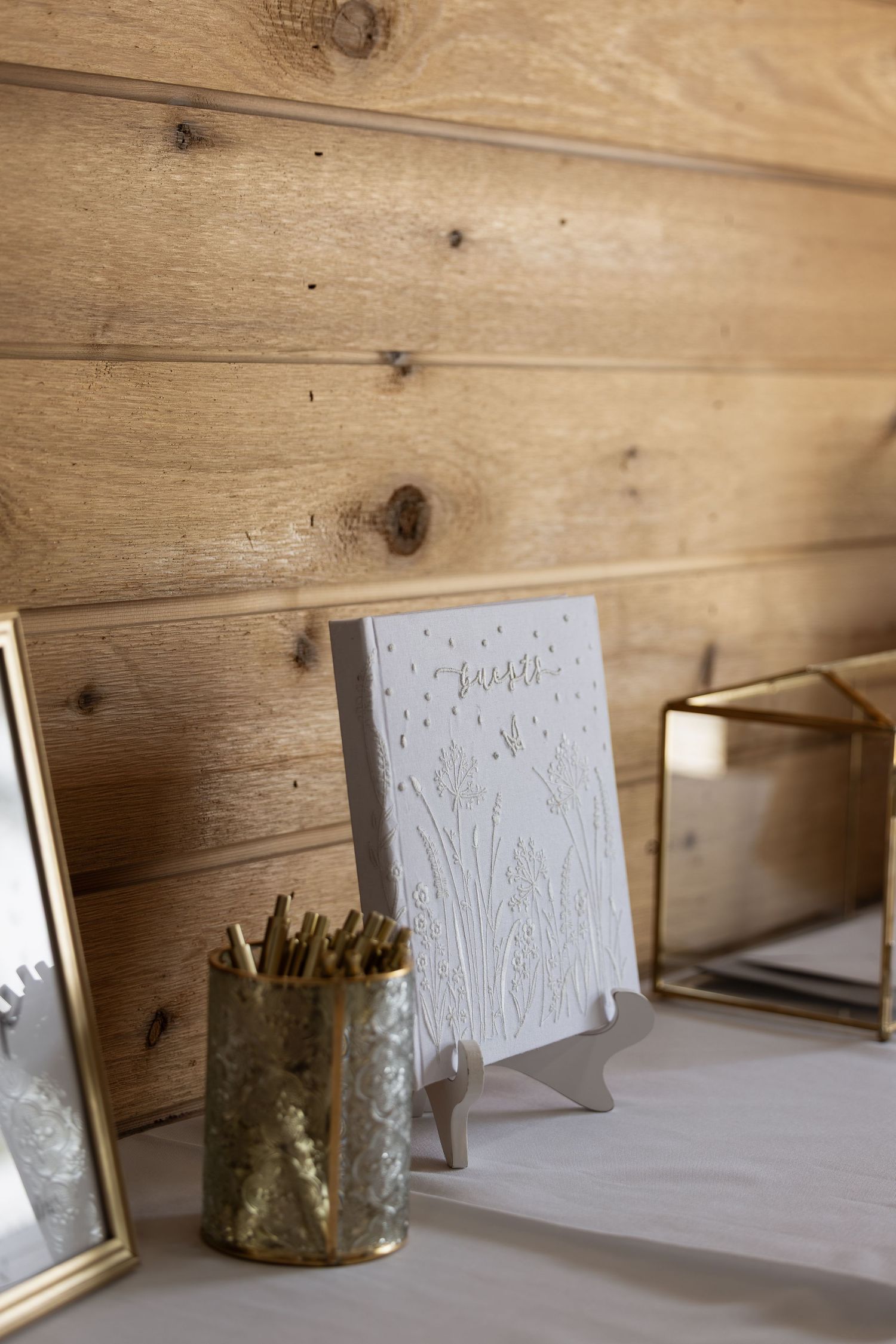 Elegant wedding guest book display with embossed white cover, gold pencil holder, and brass frames on white table.