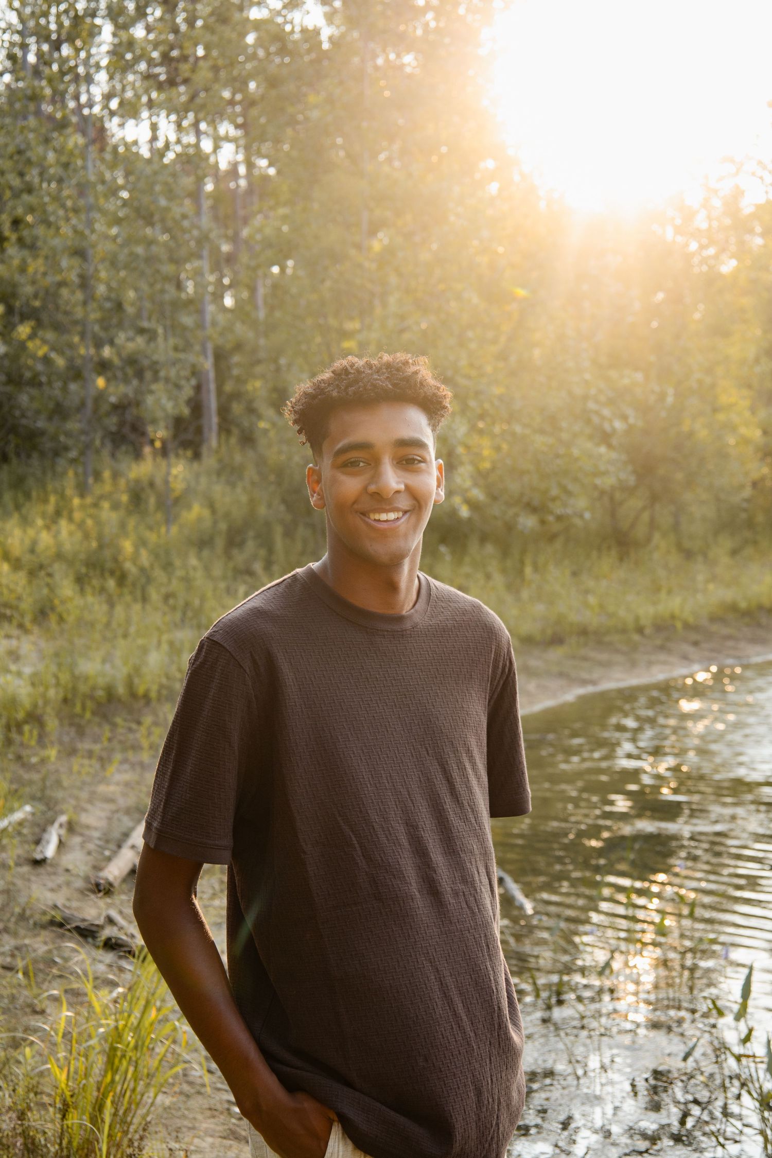 Smiling teenage boy in gray t-shirt standing by a pond in golden hour sunlight surrounded by lush green trees.