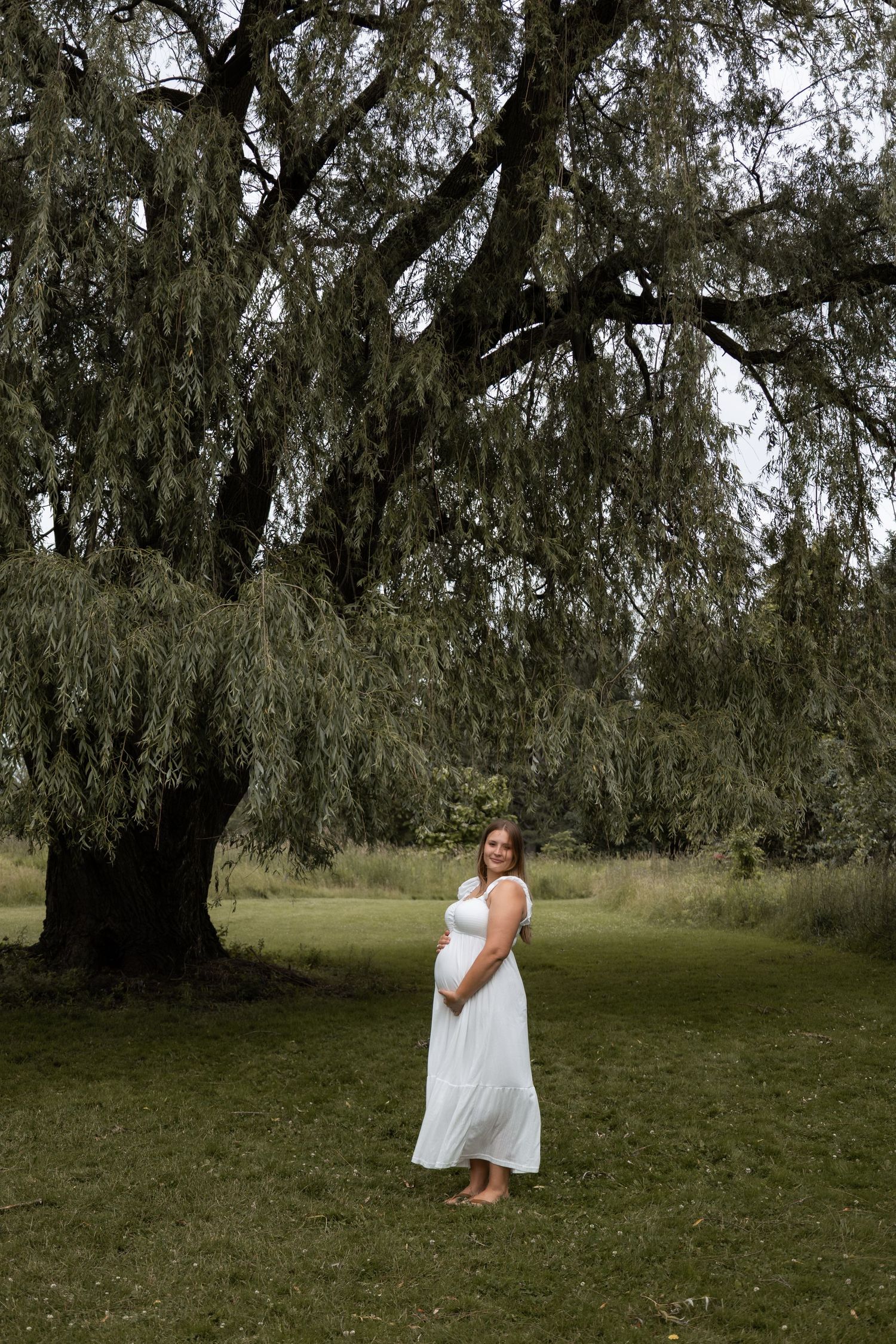 Pregnant woman in flowing white dress cradling baby bump while standing under a large weeping willow tree.
