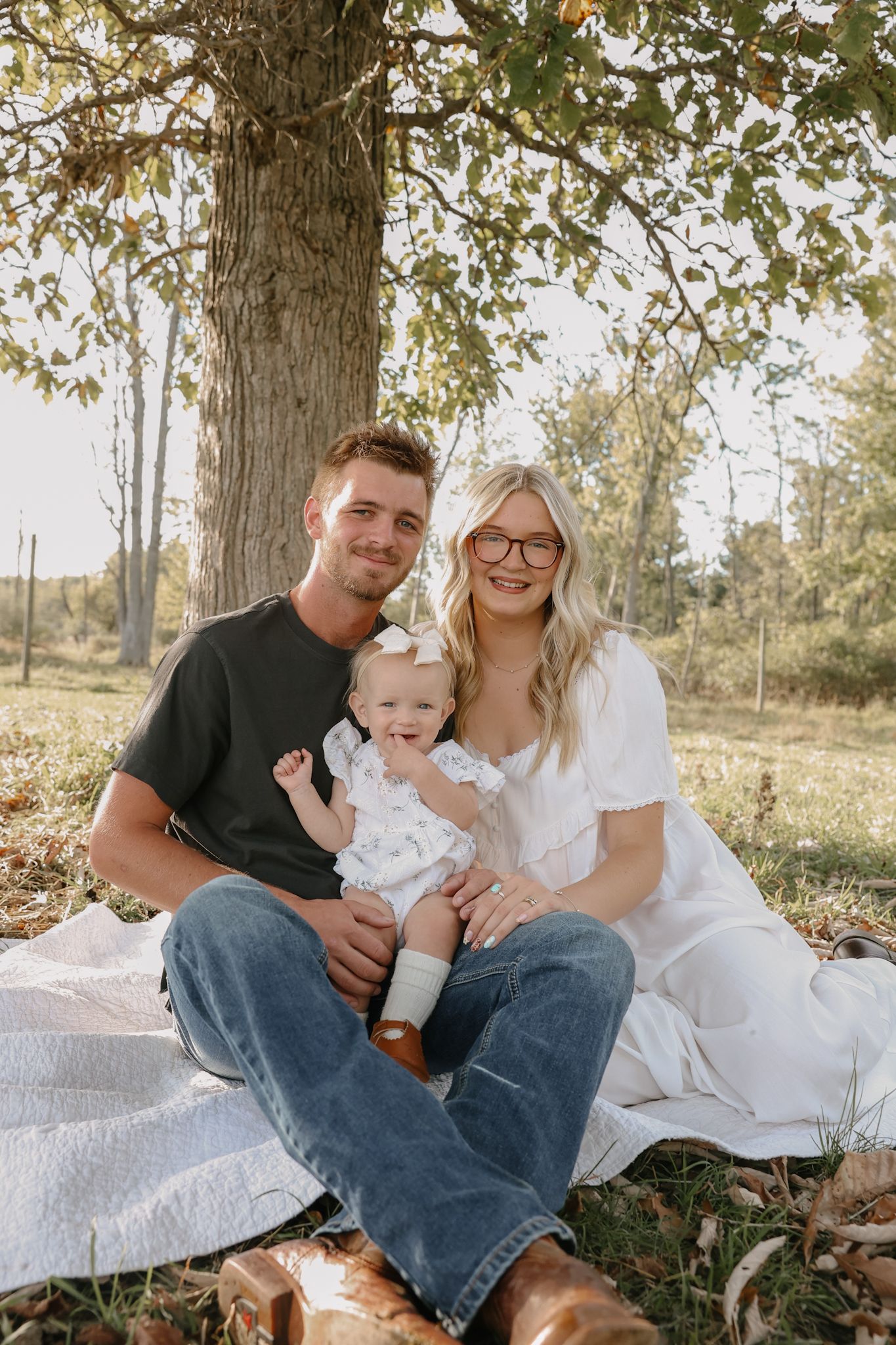 Young family with baby girl sitting on blanket under tree in golden hour outdoor photo session.
