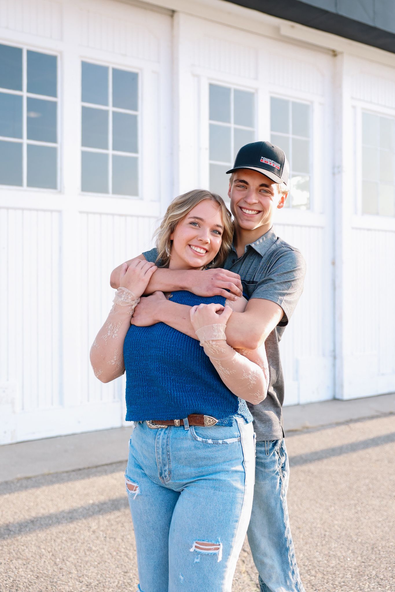 A smiling young couple embraces in front of white garage doors, the man hugging the woman from behind outdoors.