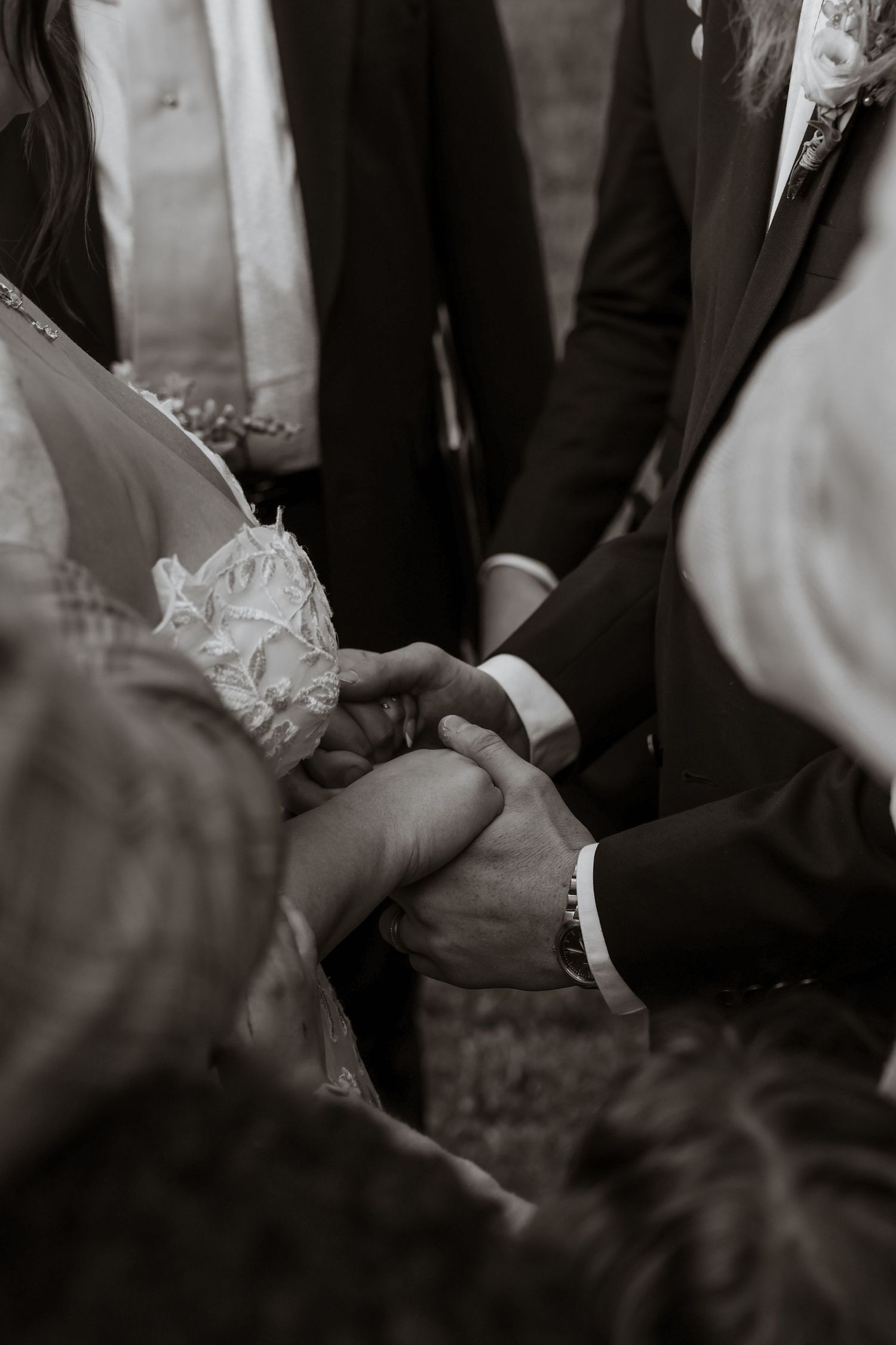 Black and white close-up of bride and groom holding hands during outdoor wedding ceremony with guests.