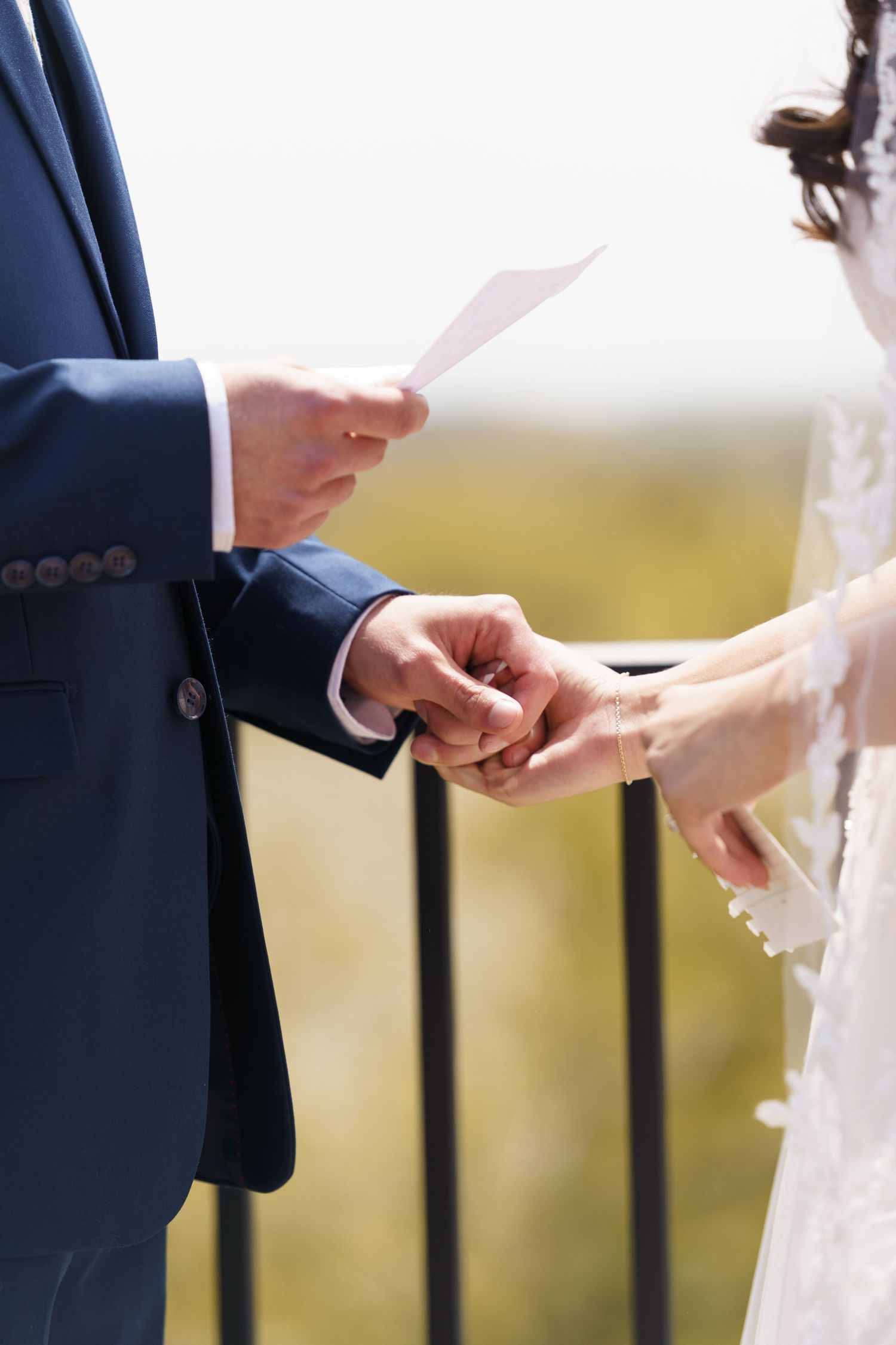 Groom in navy suit reads wedding vows while holding brides hand during outdoor ceremony.