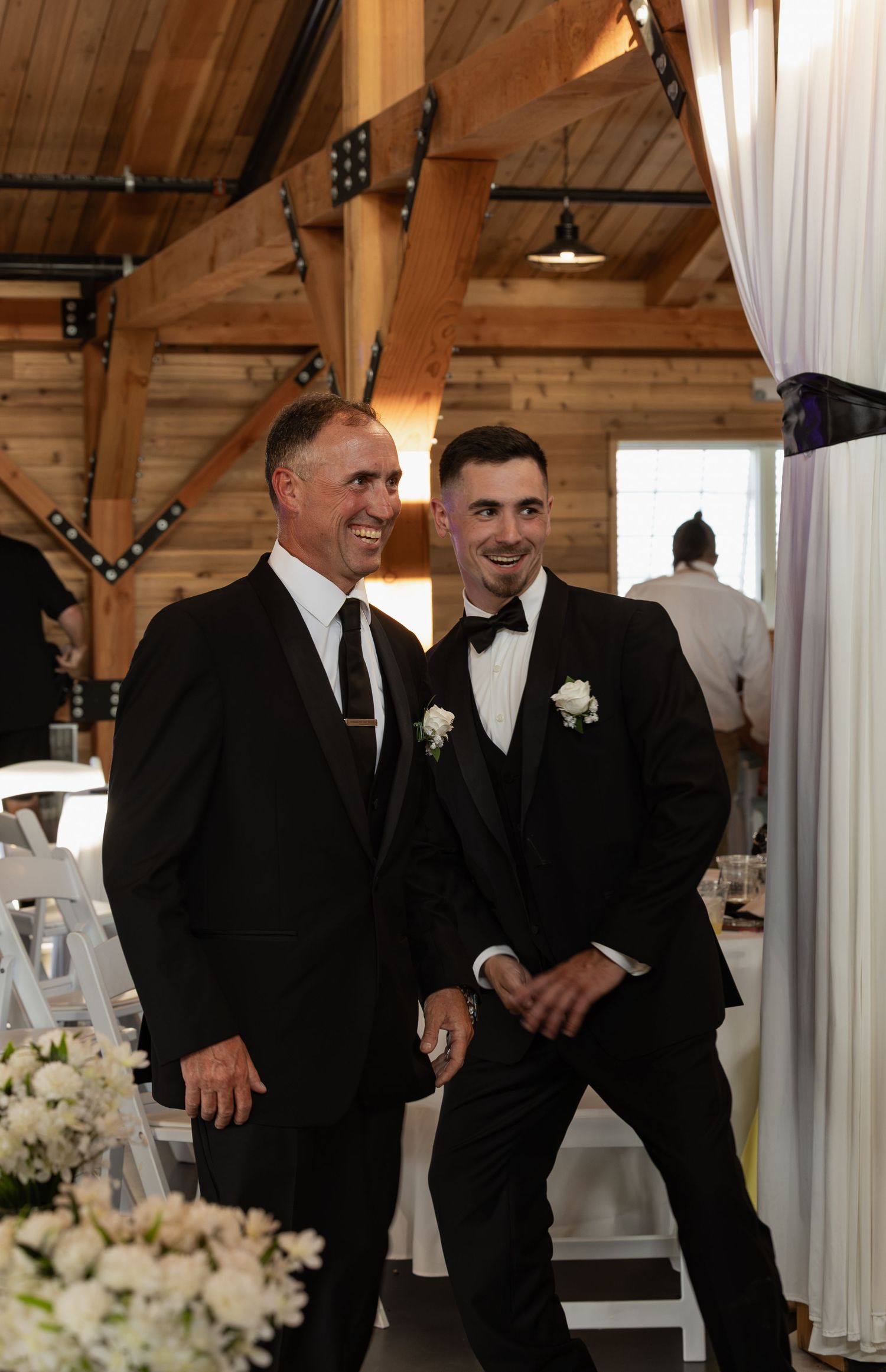 Two men in black suits smiling and walking together inside rustic barn wedding venue.
