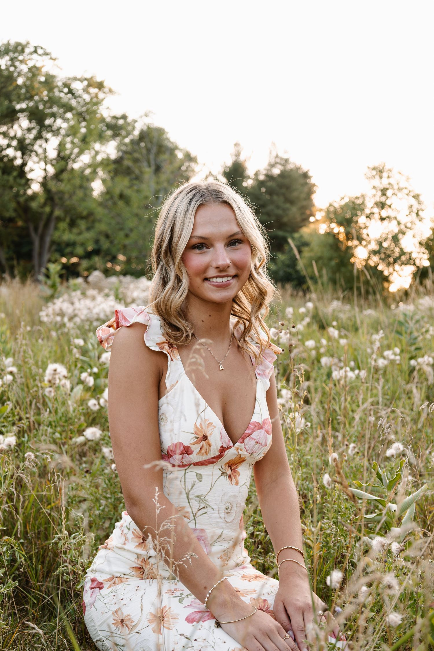 Young blonde woman in floral dress sitting in wildflower field at golden hour, smiling warmly at camera.