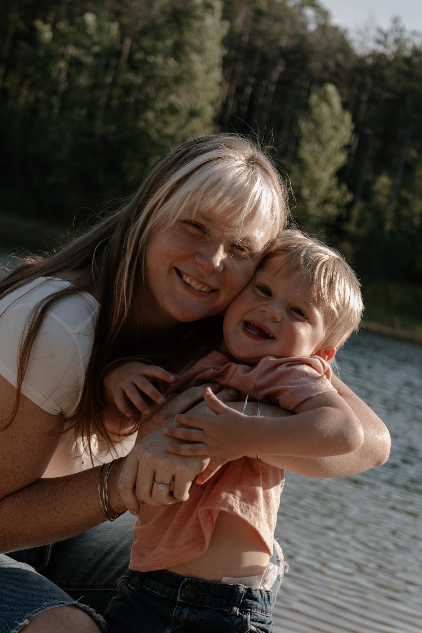 A smiling woman hugs a laughing toddler outdoors near a lake with green trees in the background.