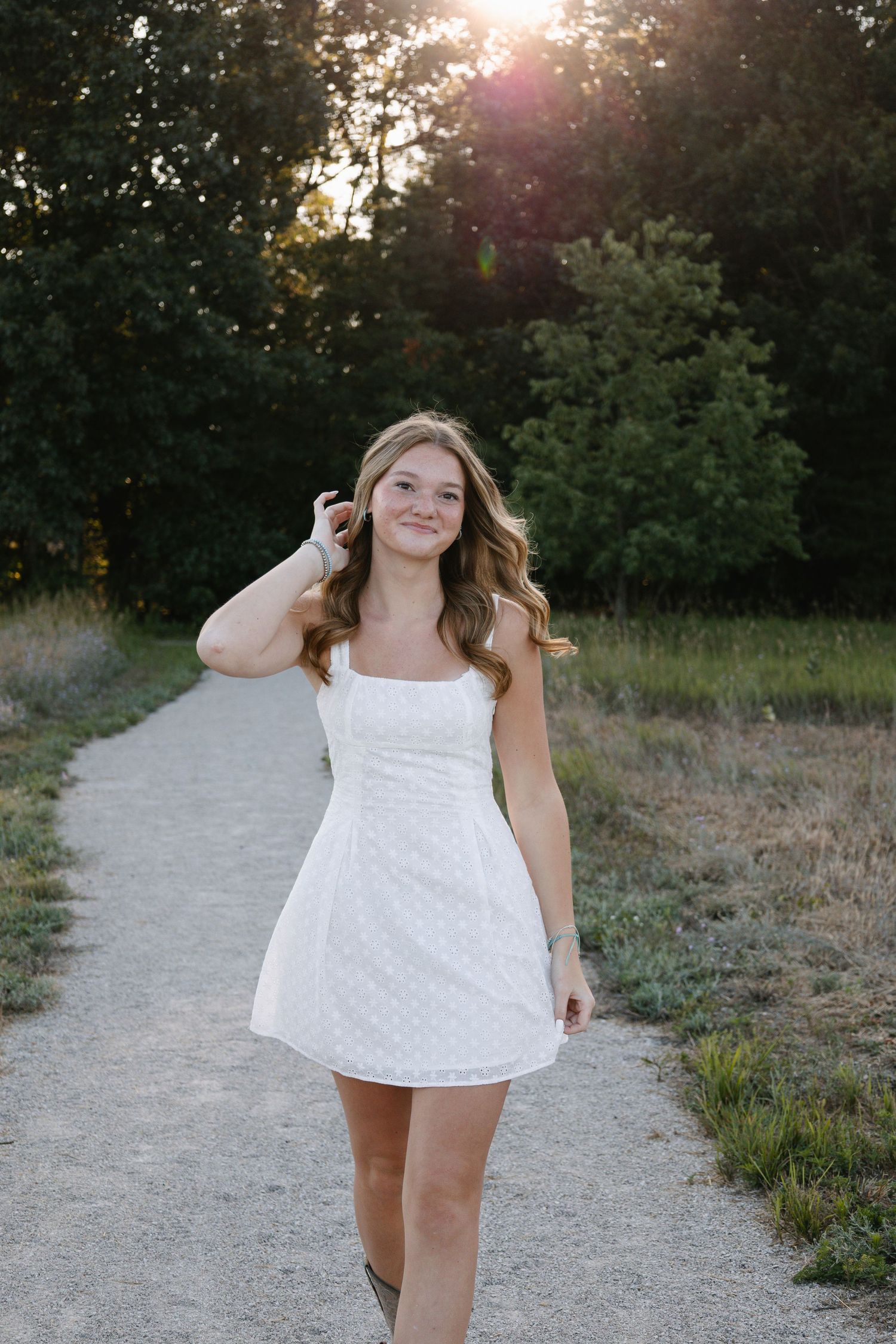 Young woman in white mini dress walking on a sunlit path surrounded by lush green trees at golden hour.