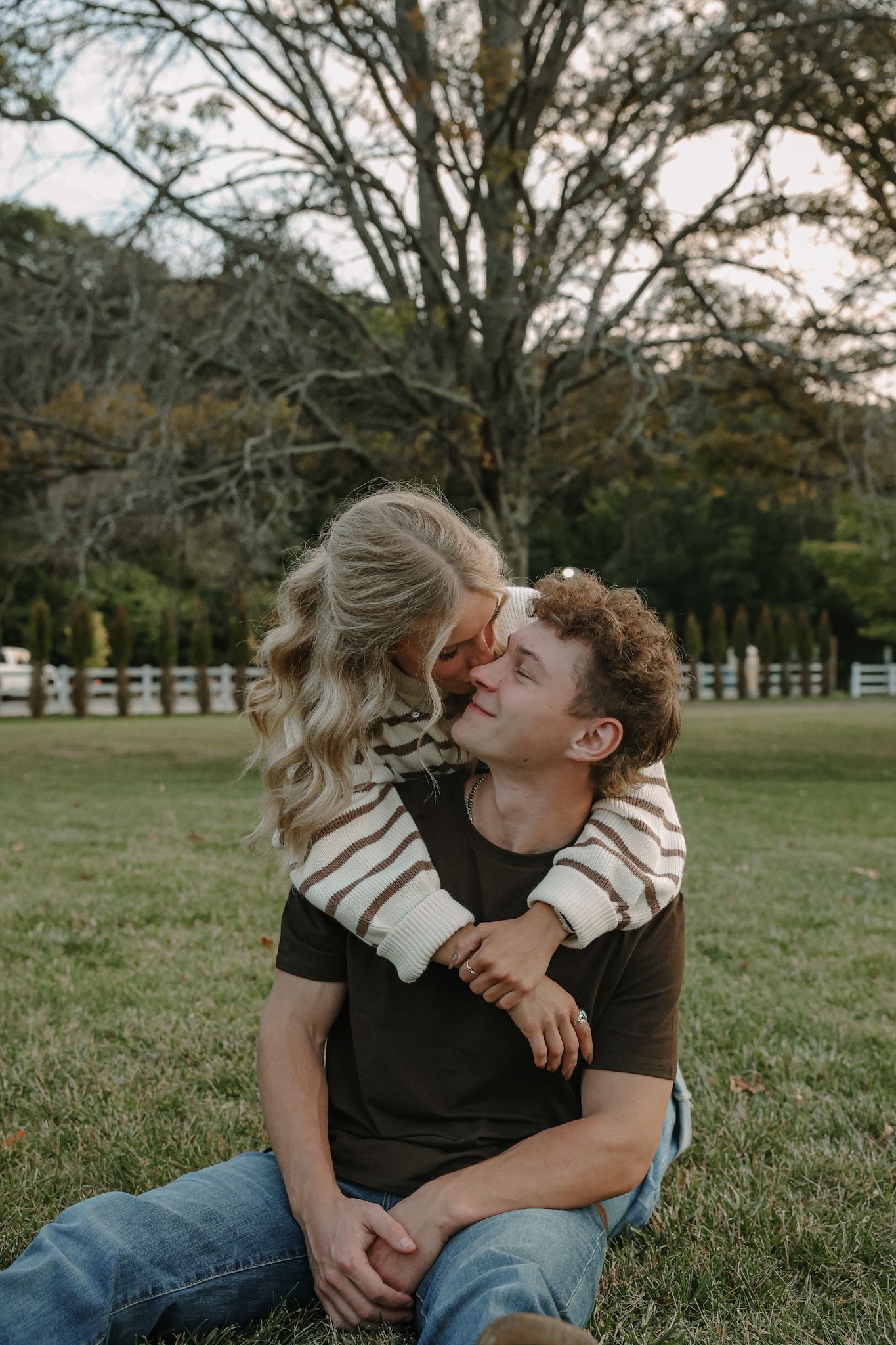 A couple shares a tender moment sitting on grass, with the woman leaning in for a kiss outdoors.