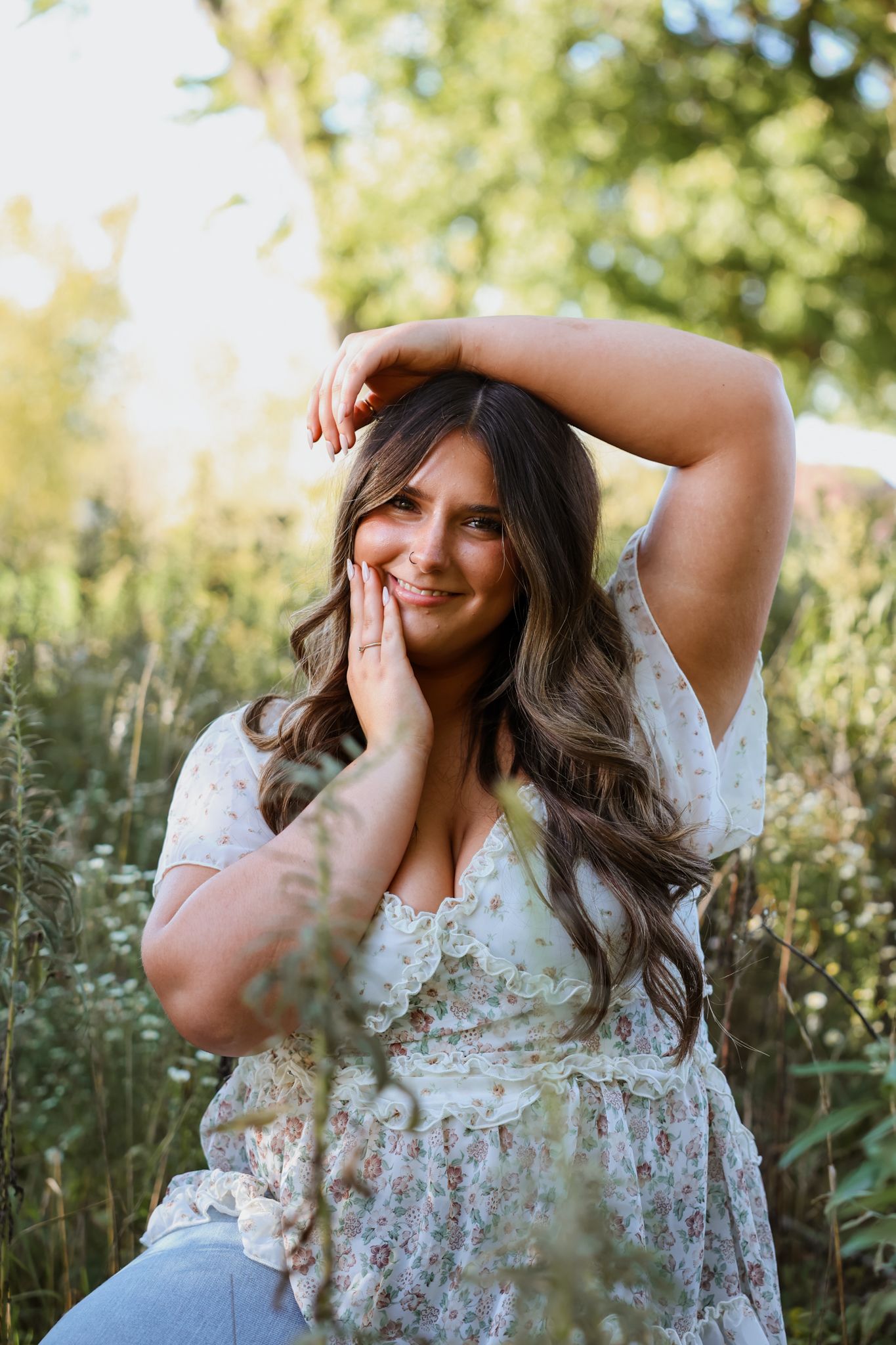 Young woman in floral dress sitting in a wildflower field, smiling with hands on her face.
