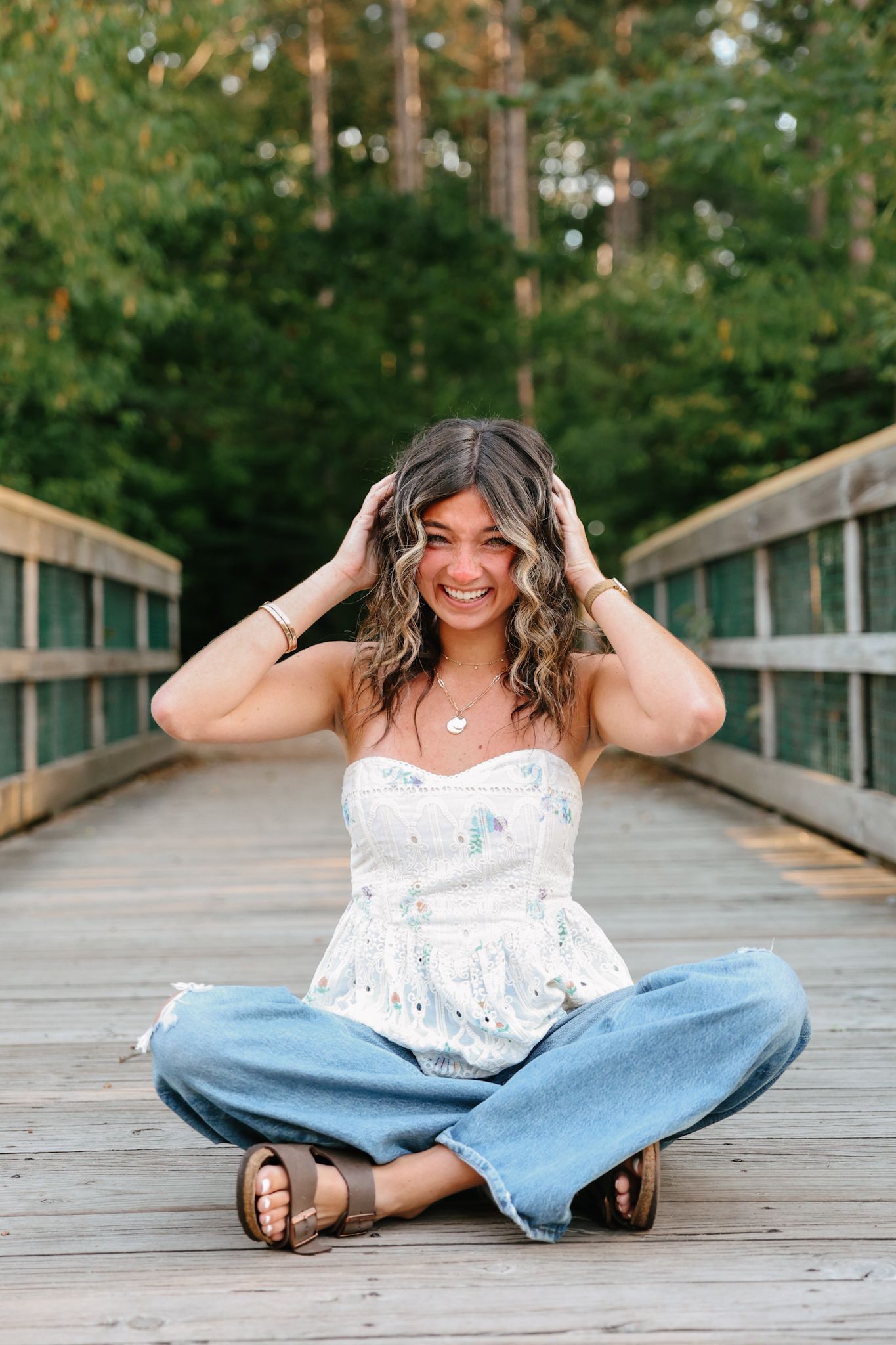 Young woman sitting cross-legged on wooden bridge laughing outdoors.