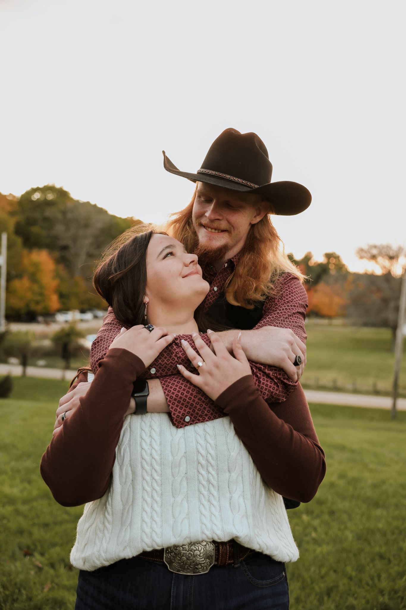 Man in cowboy hat embracing woman from behind in golden hour outdoor portrait.