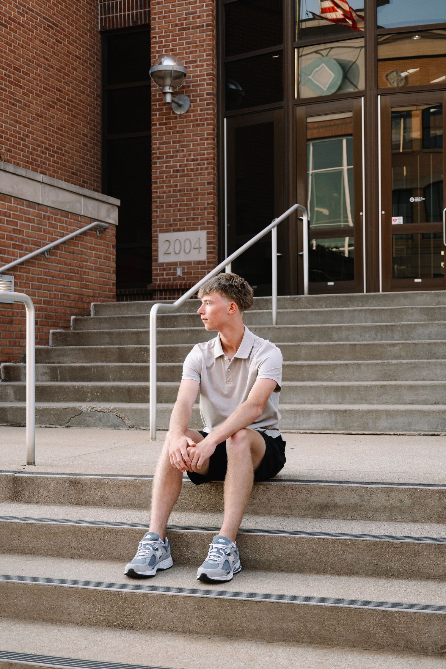 Young man sitting on brick building steps wearing polo shirt and shorts, sneakers, looking away thoughtfully.