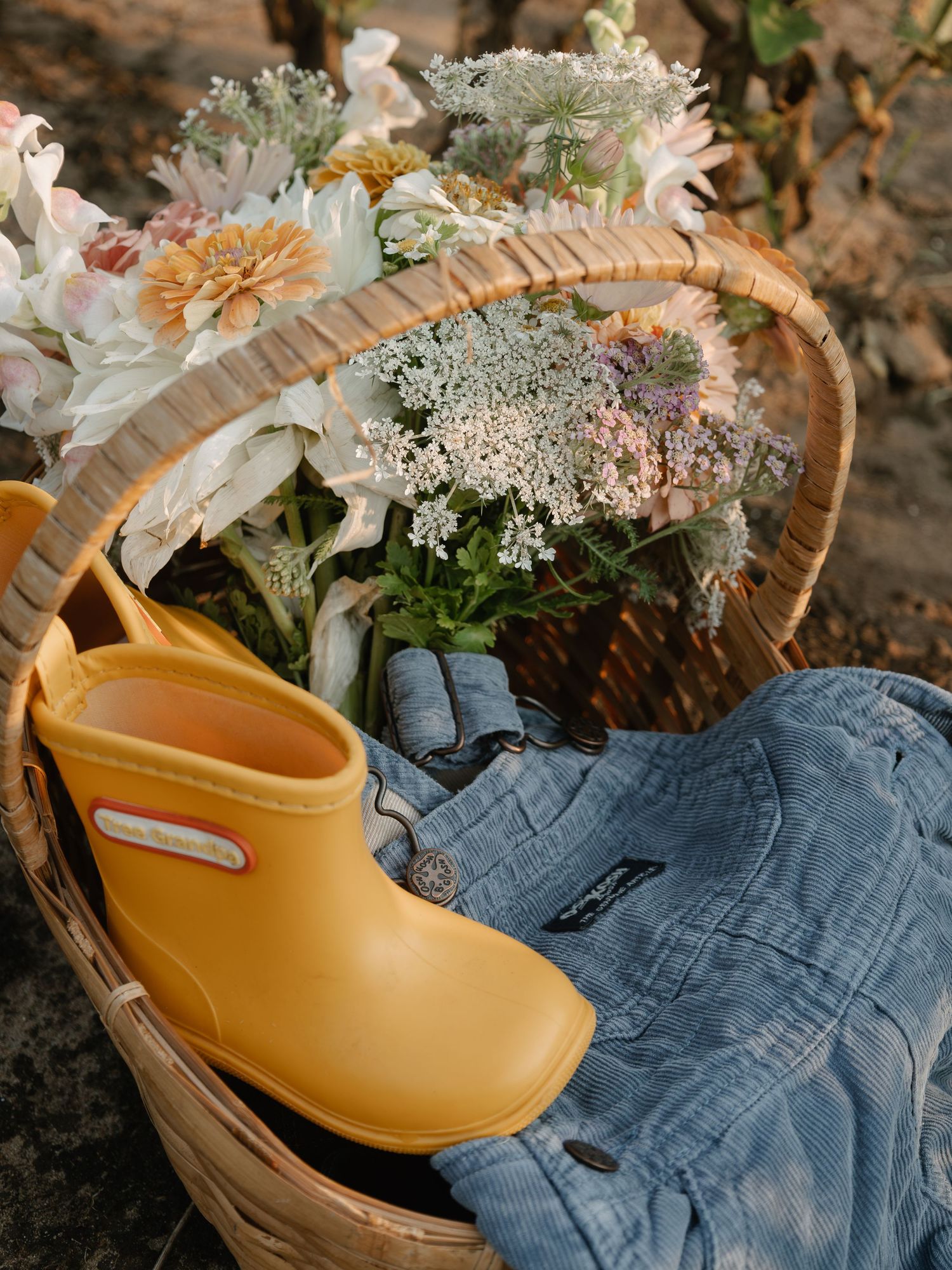 Yellow rain boot and denim overalls in a wicker basket with fresh wildflowers outdoors.