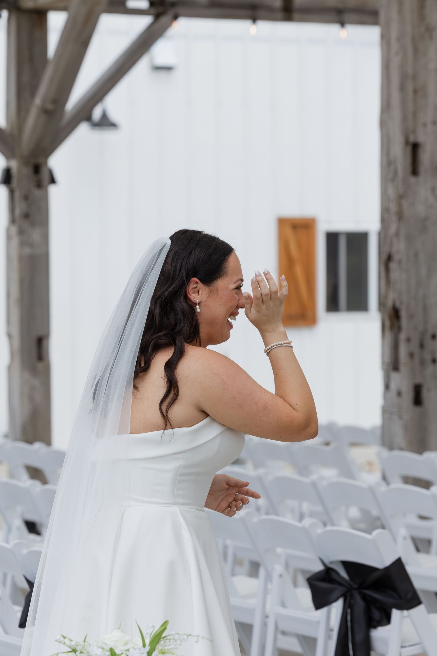 Emotional bride in white strapless gown and veil wiping tears at outdoor wedding ceremony with white chairs.