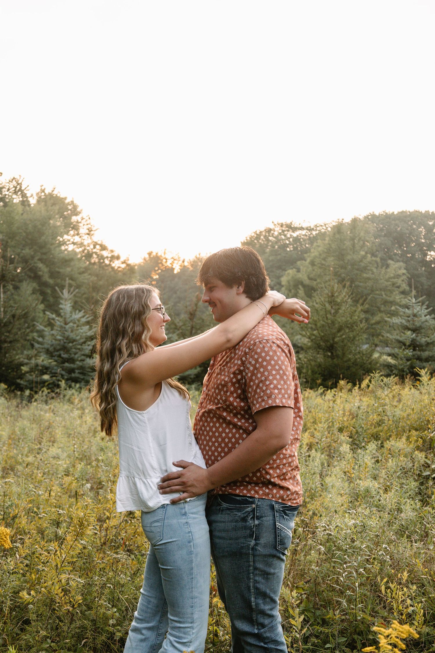Couple embracing and smiling at each other in a sunlit field with wildflowers and trees in the background.
