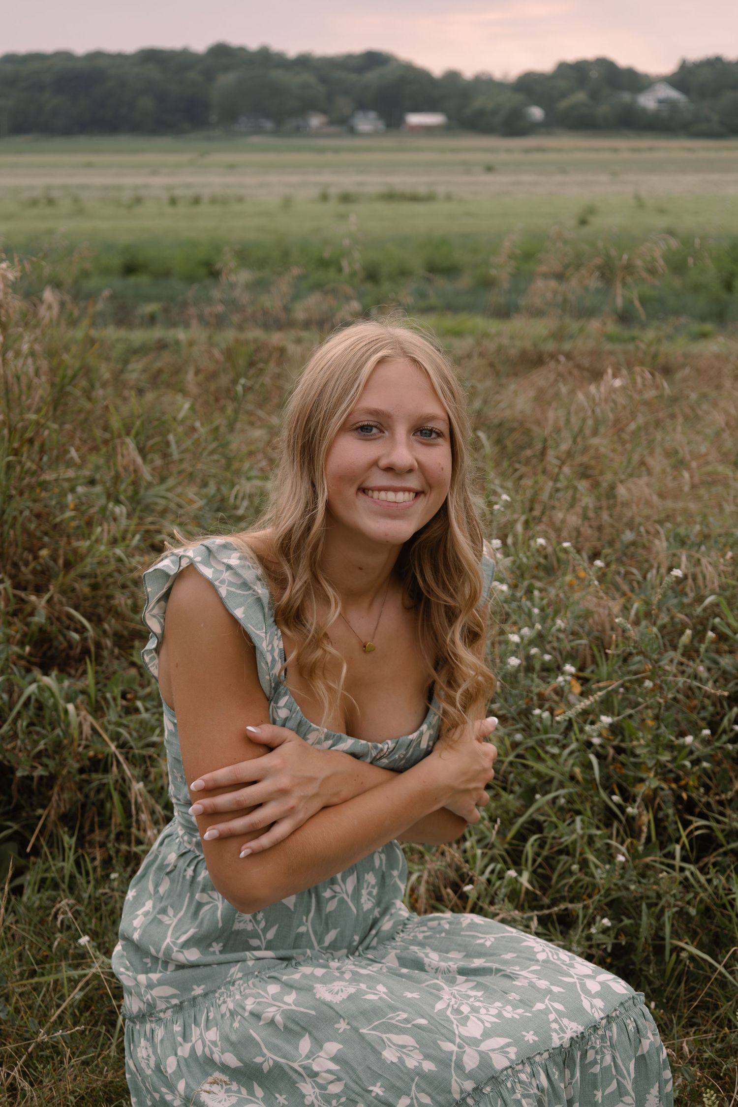 Young blonde woman in floral dress smiling while sitting in a field with green meadow backdrop.