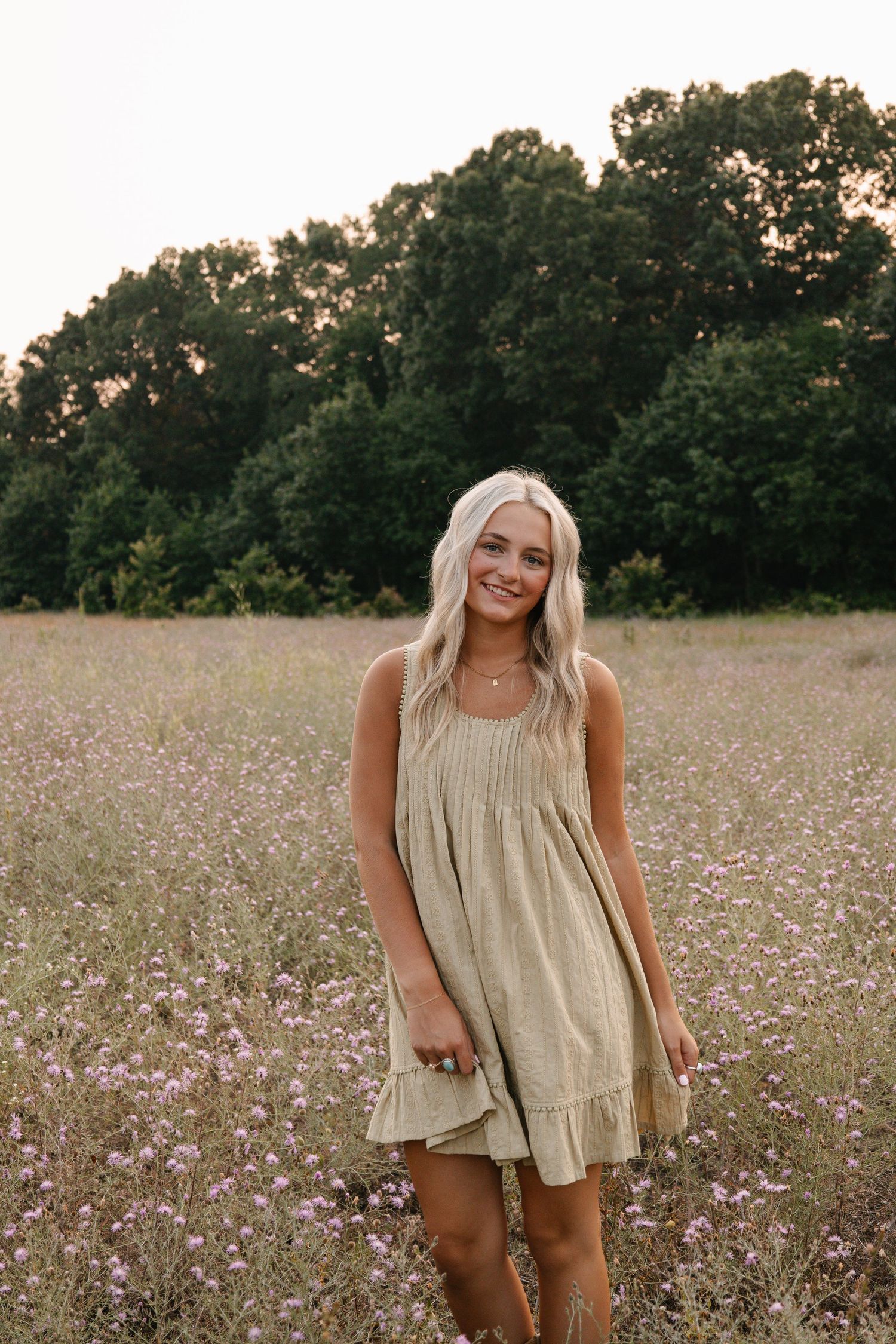 Smiling blonde woman in olive mini dress standing in wildflower field with trees in background.