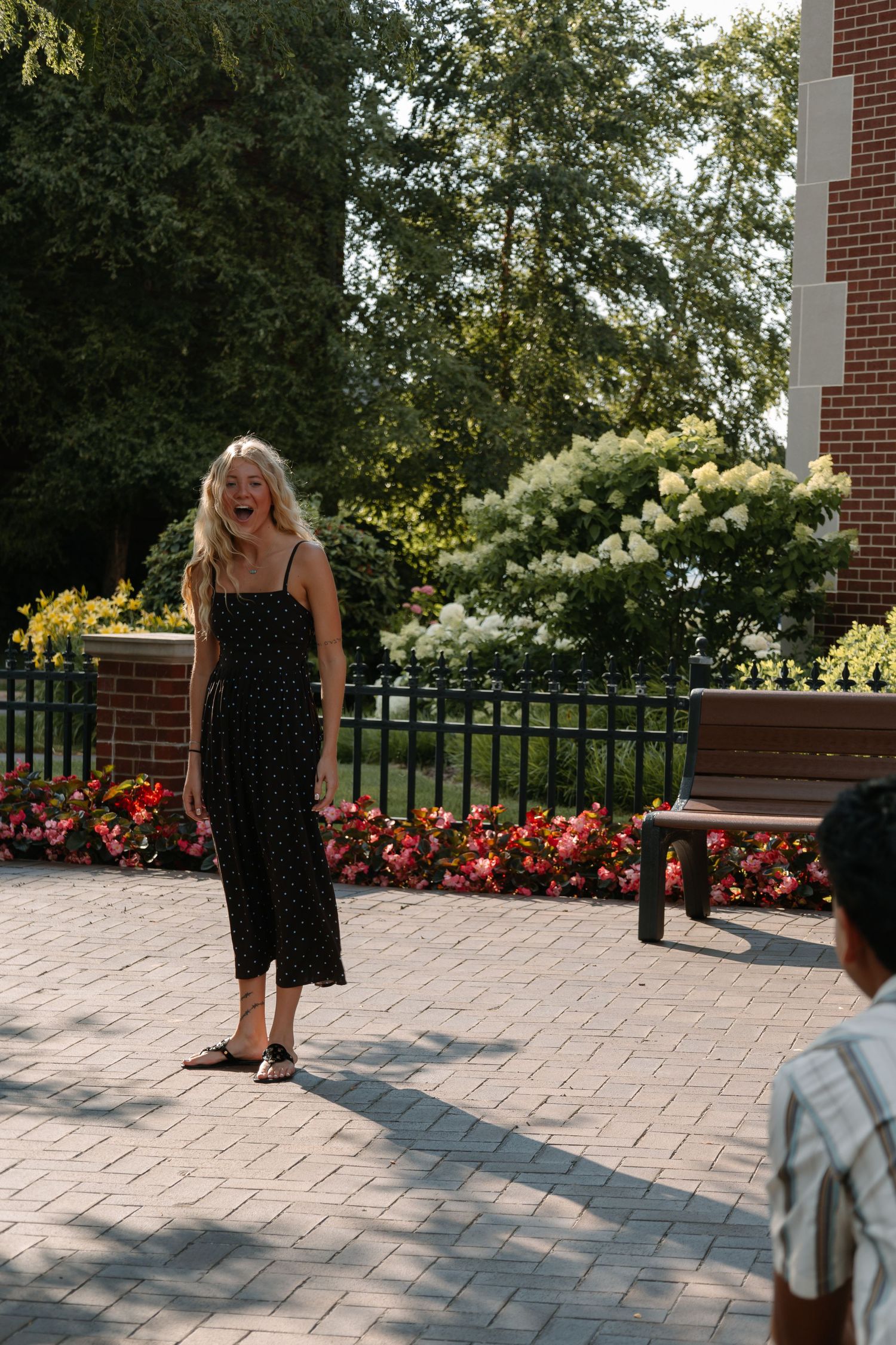 Blonde woman in black polka dot jumpsuit laughing outdoors on brick patio with colorful flowers and trees.