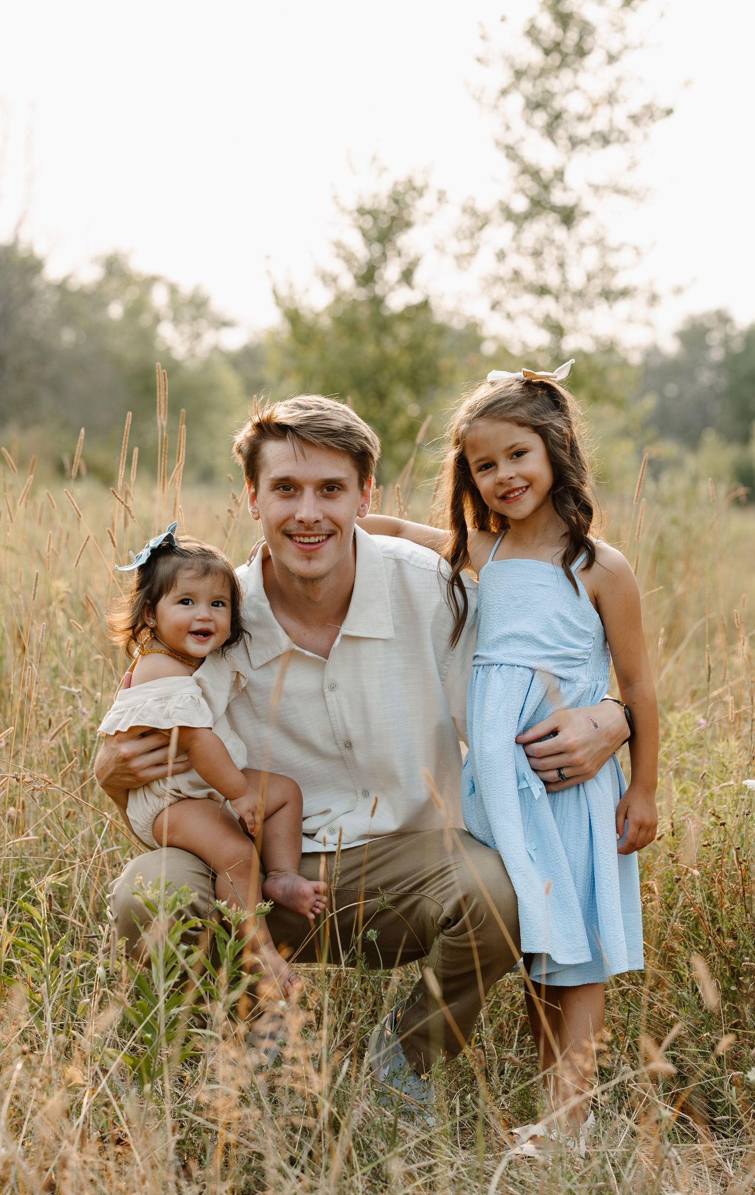 A man kneels in a golden field with two young girls, one in white and one in a blue dress, smiling outdoors.