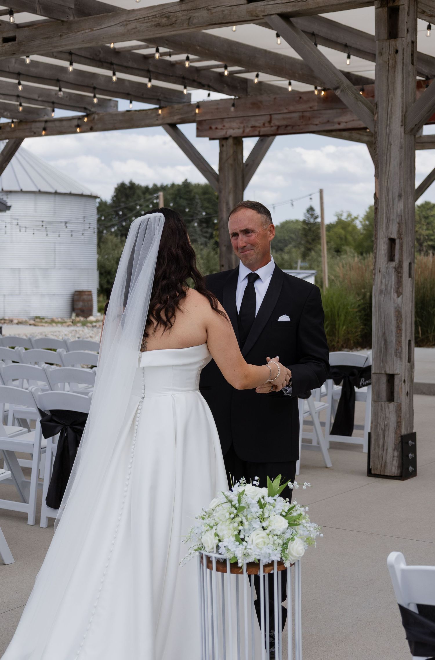 Bride in white strapless gown holding white bouquet faces groom in black suit at rustic outdoor wedding ceremony.