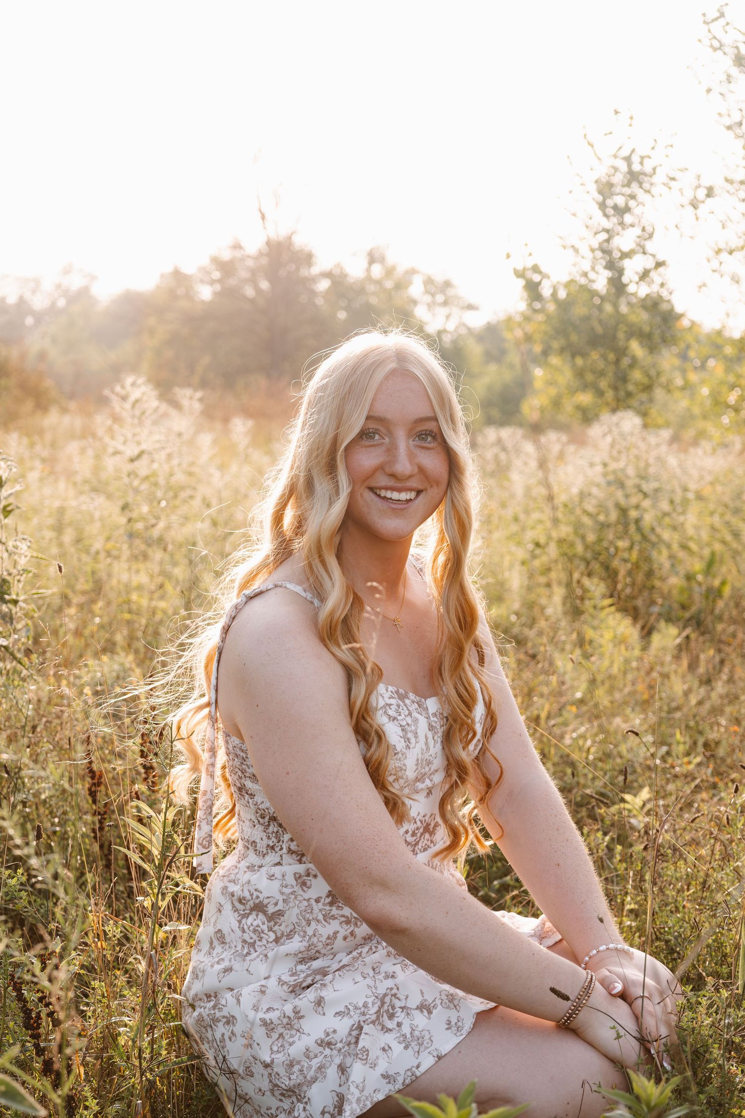 Young woman with long blonde hair in floral dress sitting in golden sunlit field, smiling warmly at camera.
