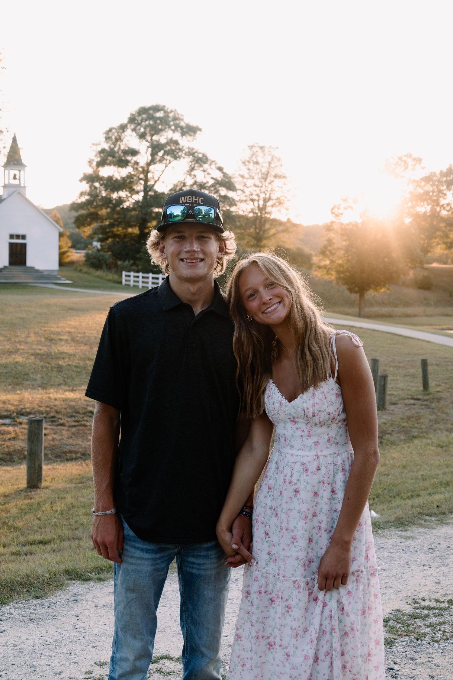 Young couple holding hands smiling at sunset near a white country church on a rural path.