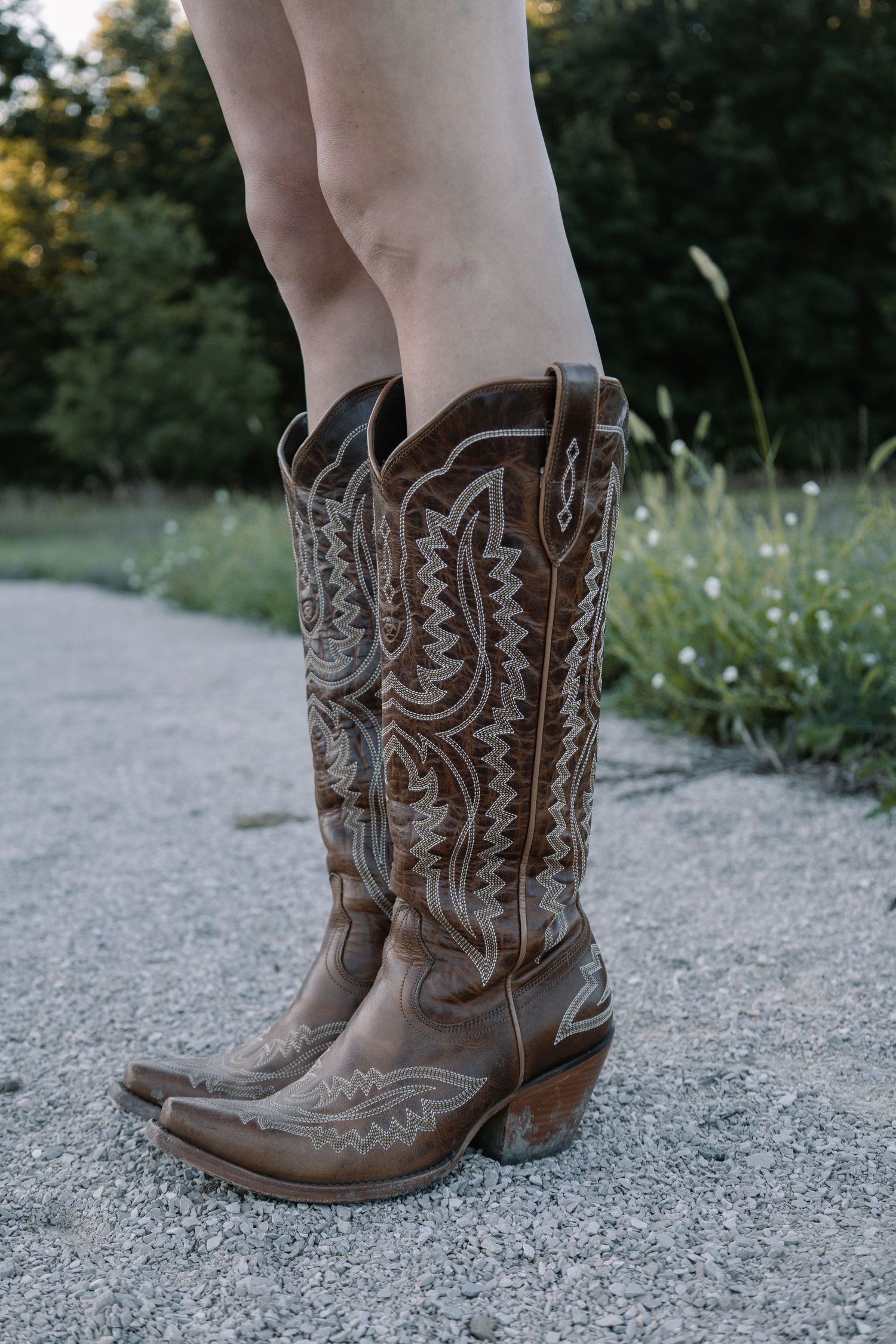Brown leather cowboy boots with white stitching worn outdoors on gravel path with greenery background.