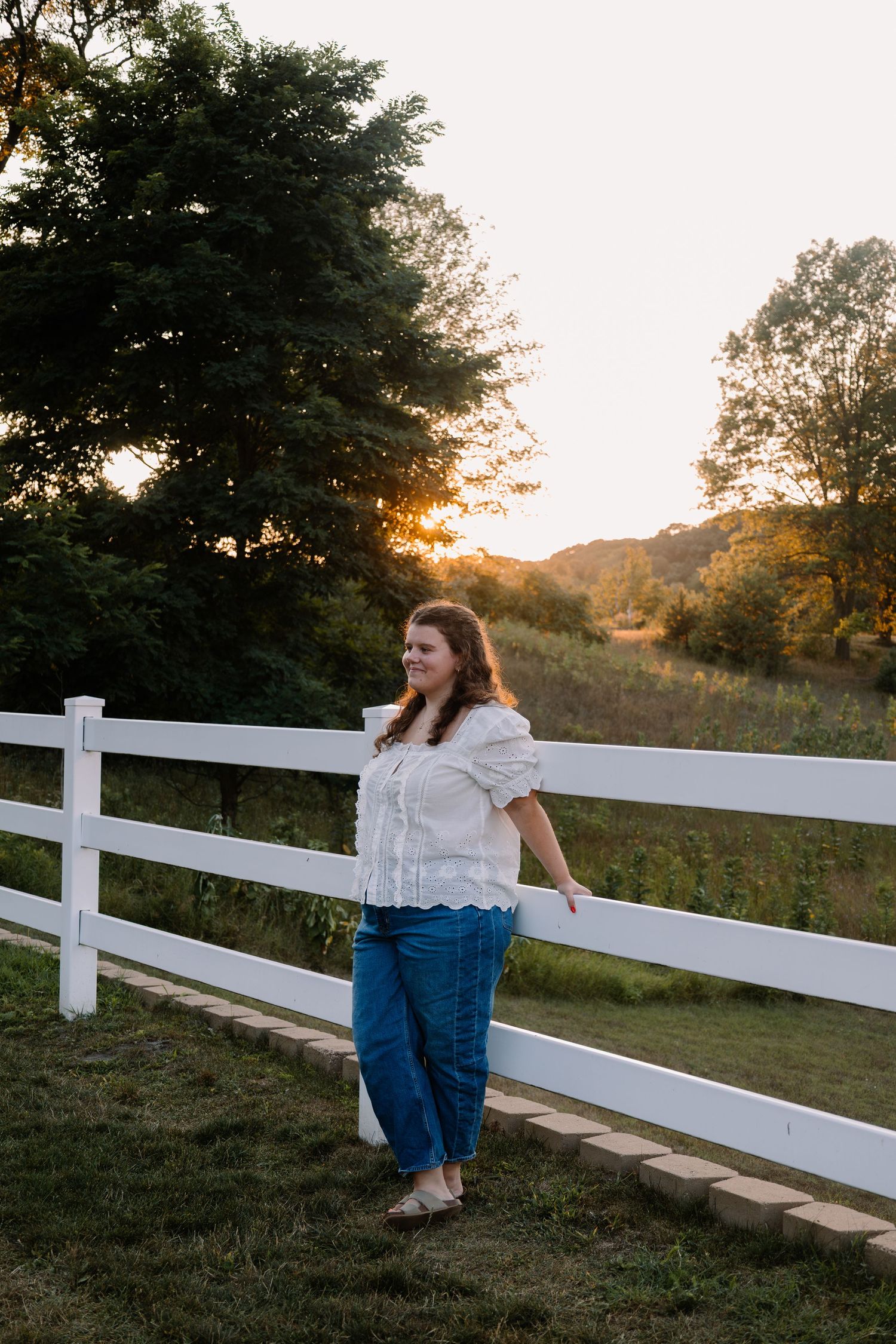 Woman in white top and jeans leaning on white fence at golden hour sunset with lush green trees.