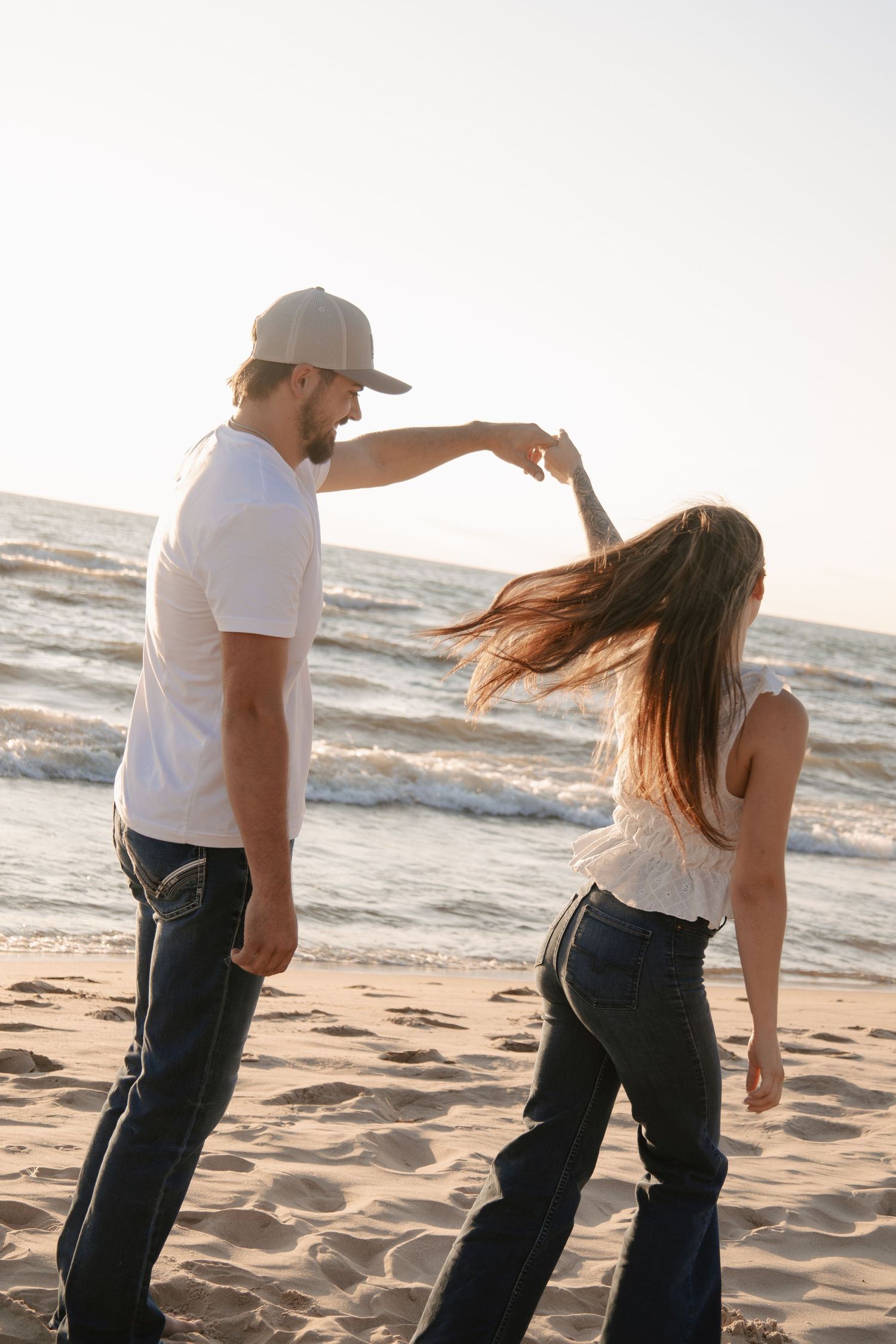 Couple dancing on sandy beach at sunset, man in white tee and cap spinning woman in jeans by the ocean.