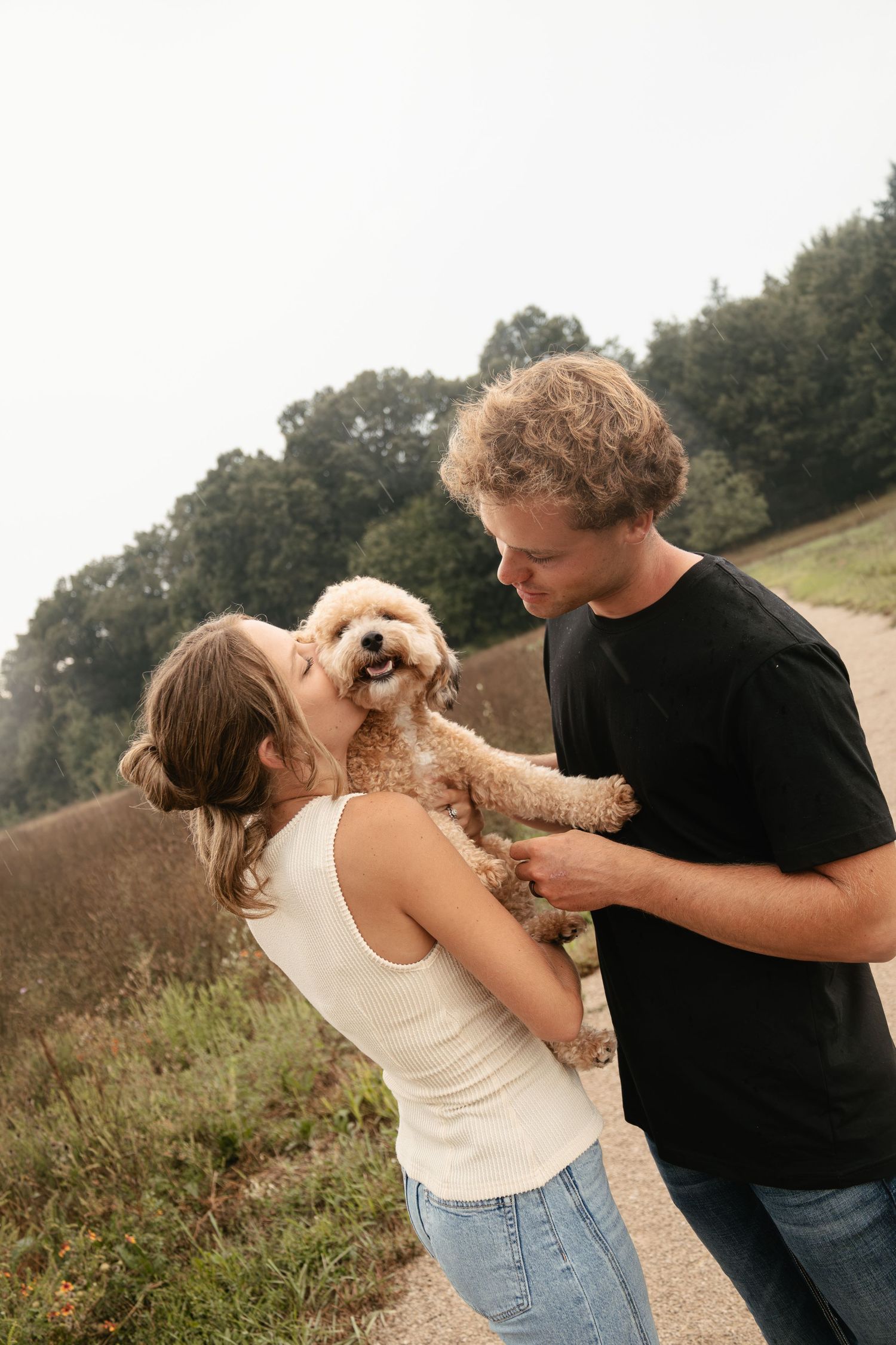 Couple sharing a sweet moment kissing their fluffy golden doodle puppy outdoors on a scenic dirt path.