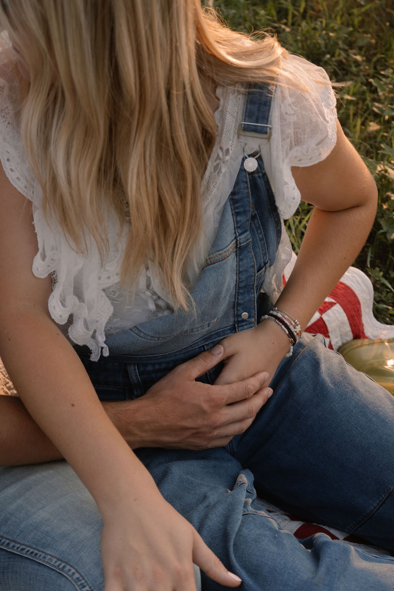 Close-up of woman in denim overalls and white lace top sitting in grass at sunset with hand resting on knee.