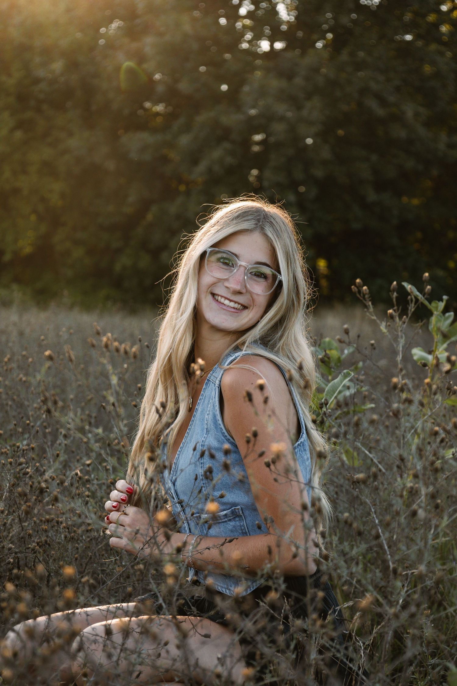 Smiling blonde teen girl with glasses sits in a sunlit field wearing a denim romper, surrounded by wildflowers.