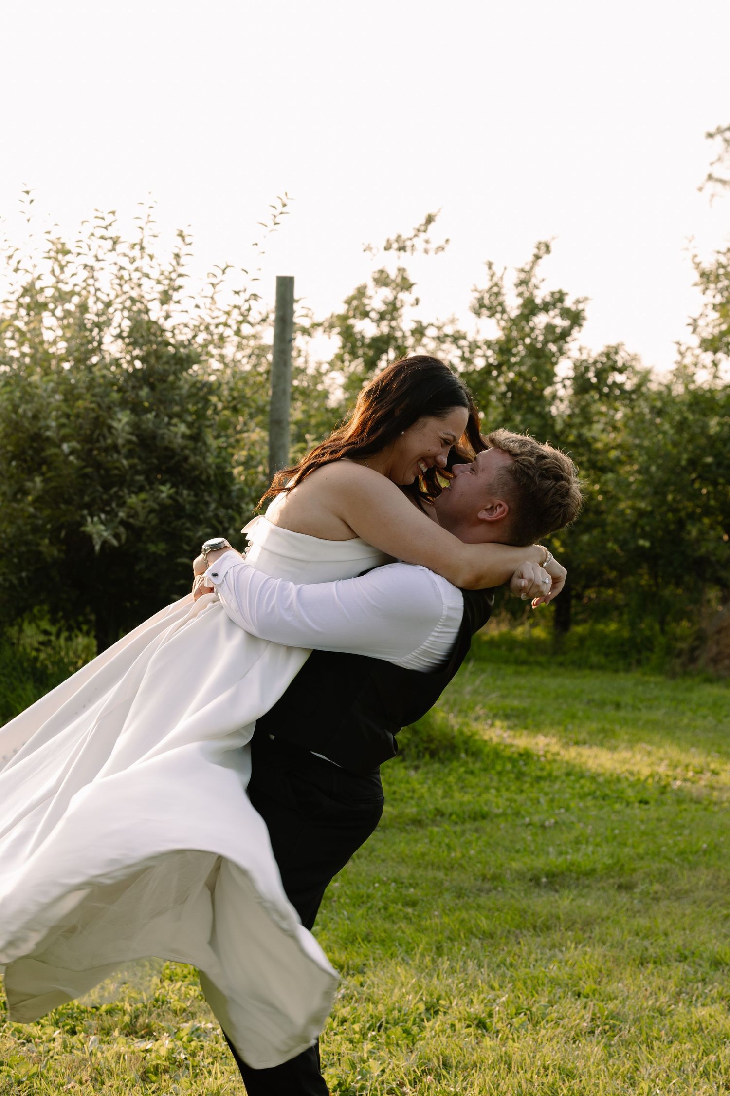 Groom lifts bride in white dress outdoors at golden hour in lush green orchard setting.