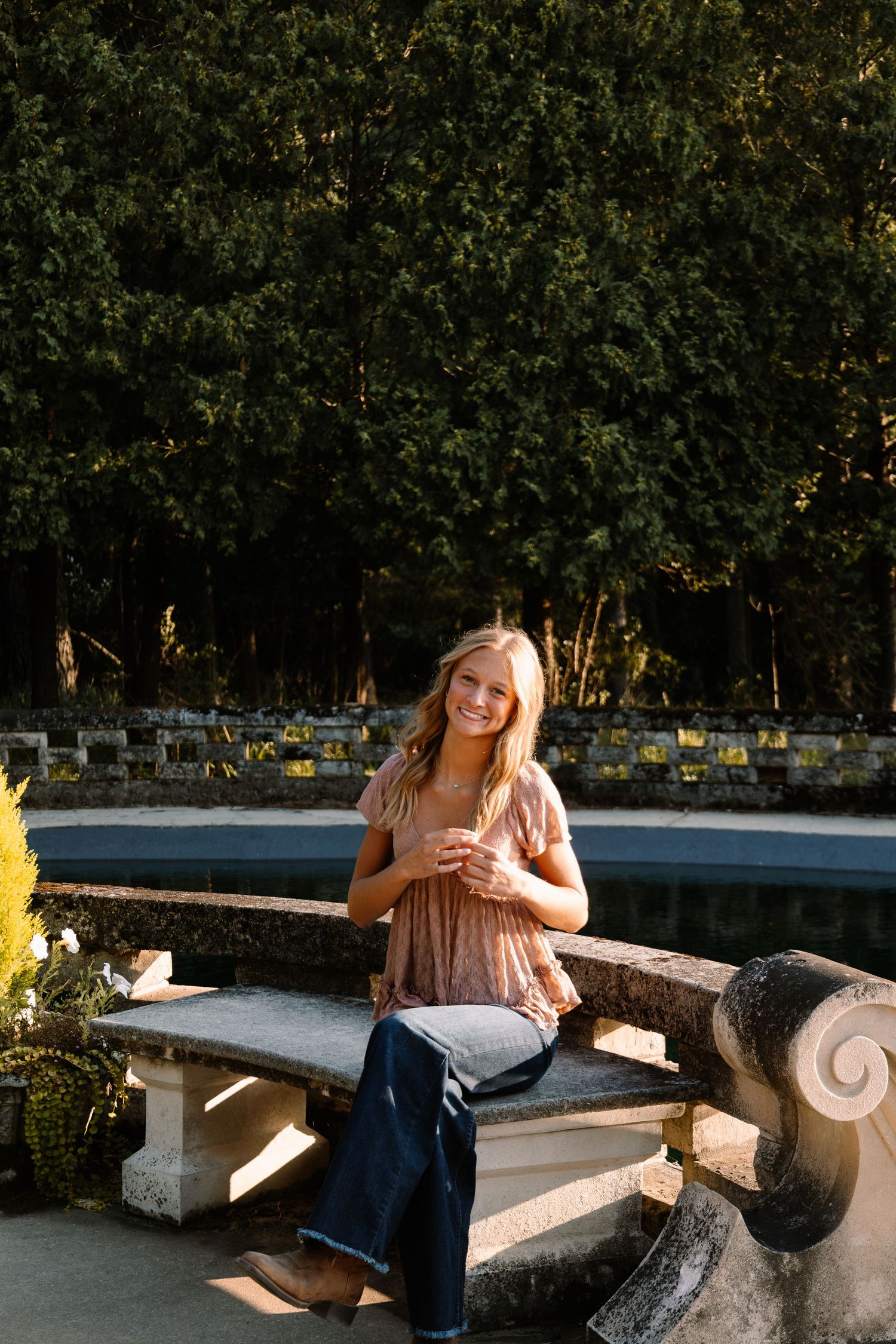 Smiling blonde woman sitting on a stone bench by a garden fountain in golden hour light, wearing a pink top and jeans.