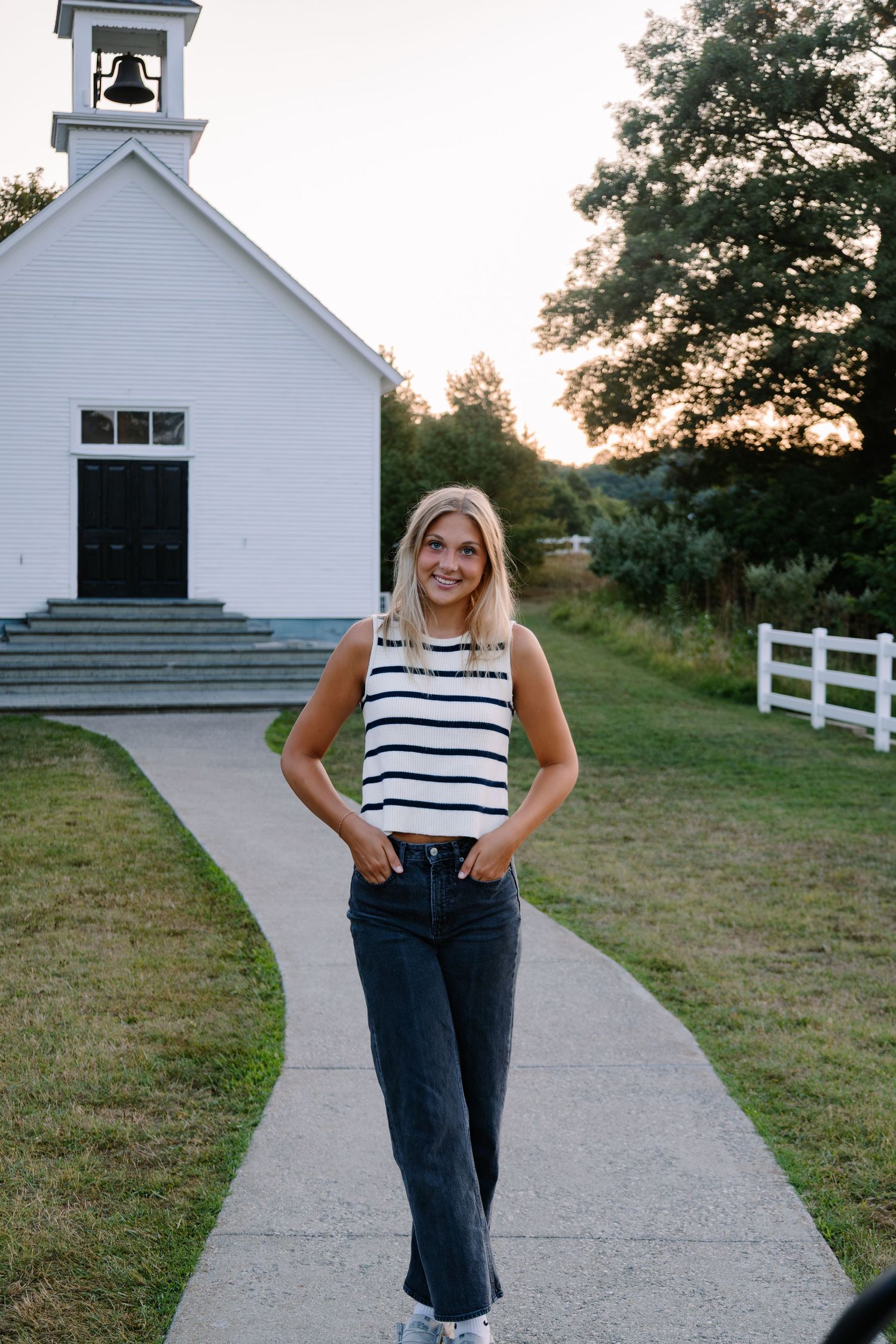 Smiling blonde woman in striped top and dark jeans stands on a pathway in front of a white chapel at sunset.