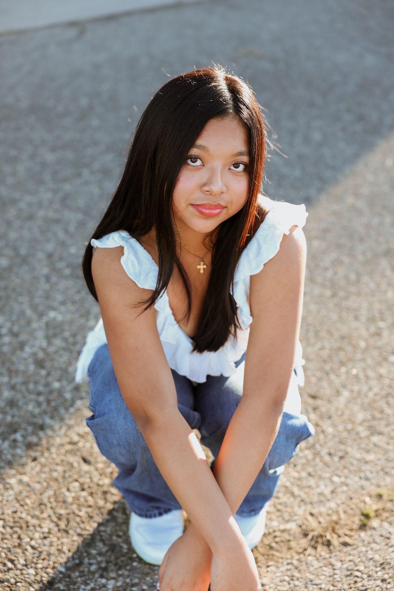 Young Asian teen in white ruffle top and jeans crouches on pavement in bright sunlight.