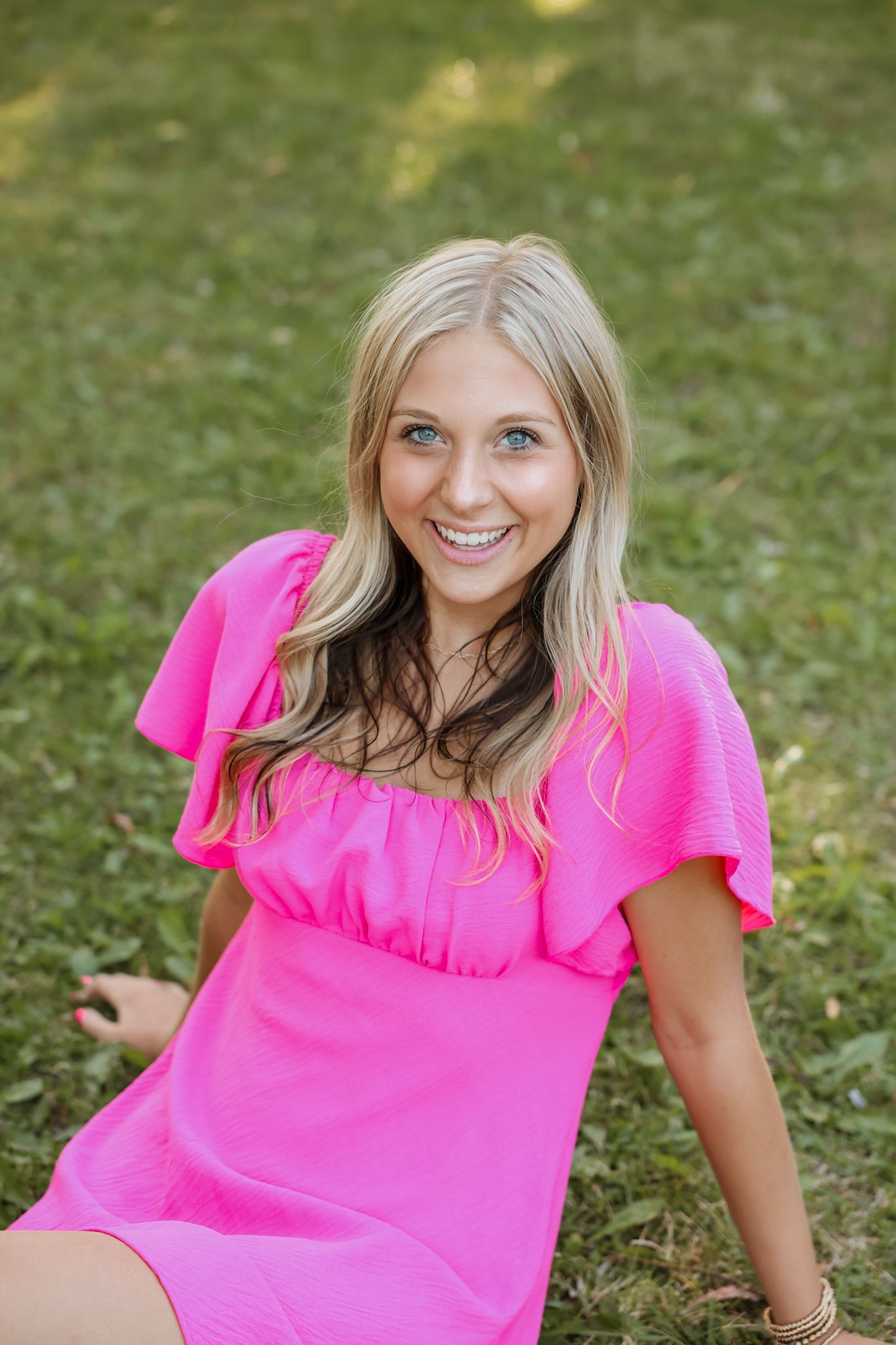 Smiling blonde woman in hot pink dress sitting on green grass in an outdoor park setting.