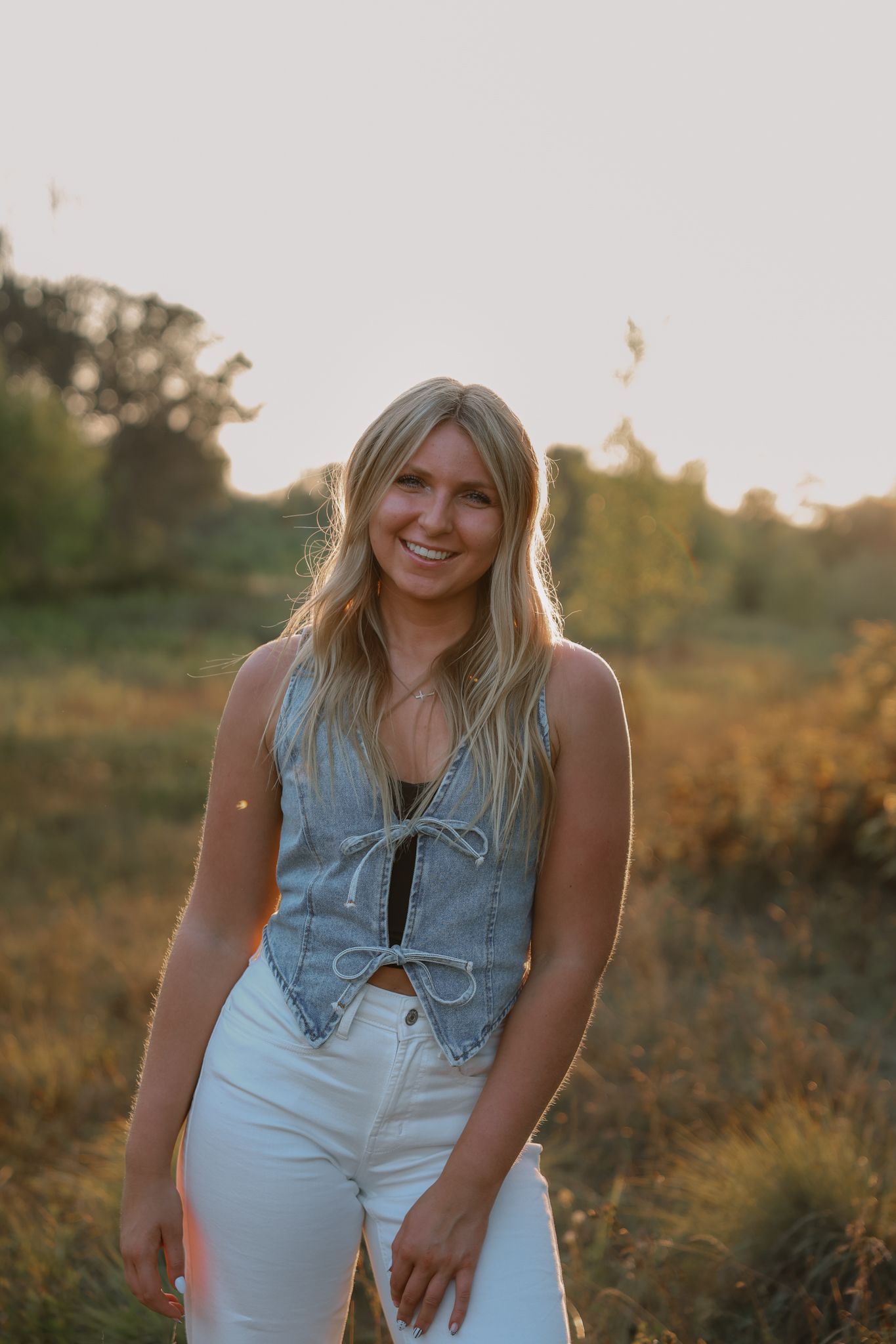Smiling blonde woman in denim vest and white jeans posing in a golden field during a warm sunset photo session.