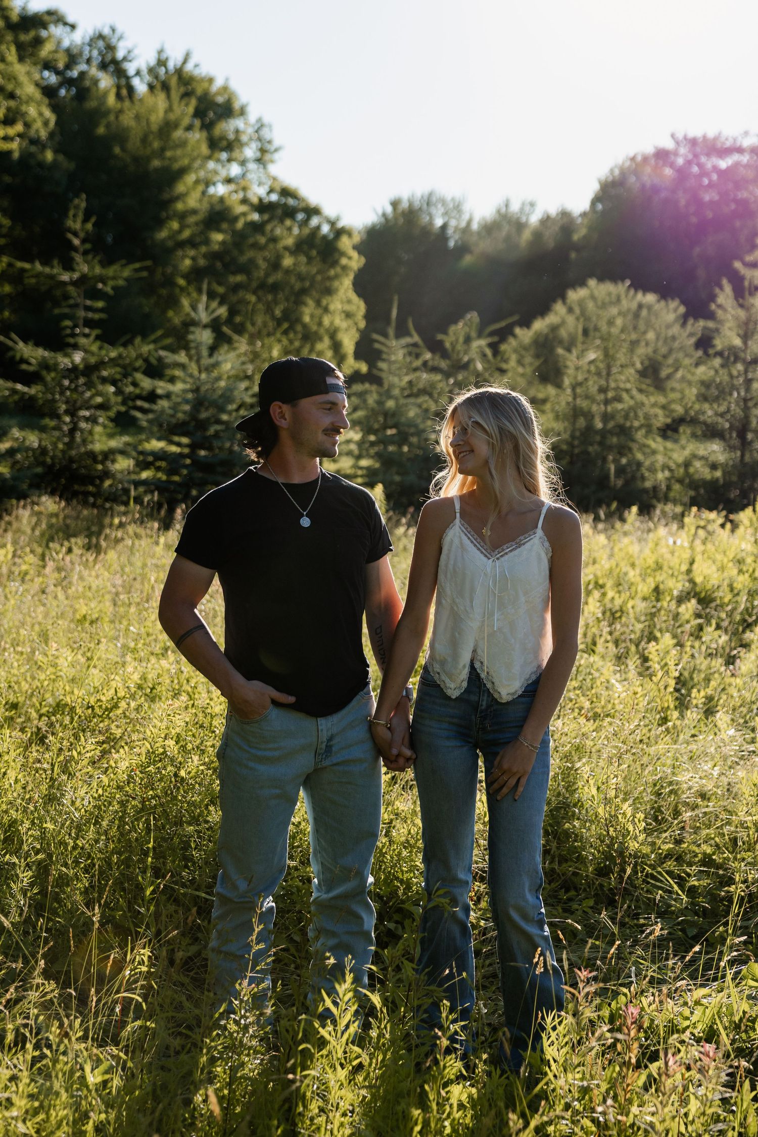 Couple holding hands and smiling at each other in a sunlit green meadow, man in black tee and cap, woman in white top.