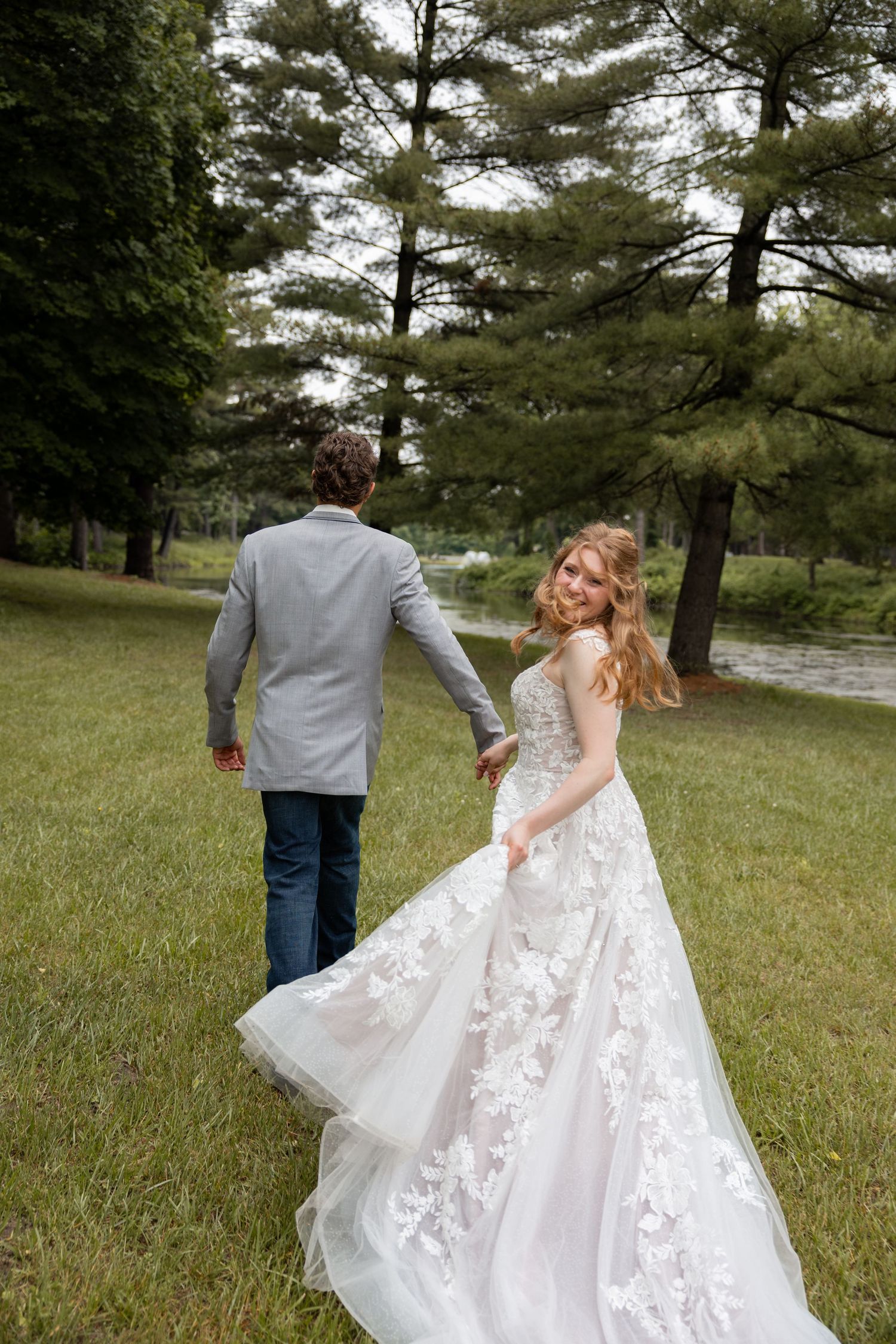 Bride in floral lace gown glancing back while holding groom's hand walking through park.