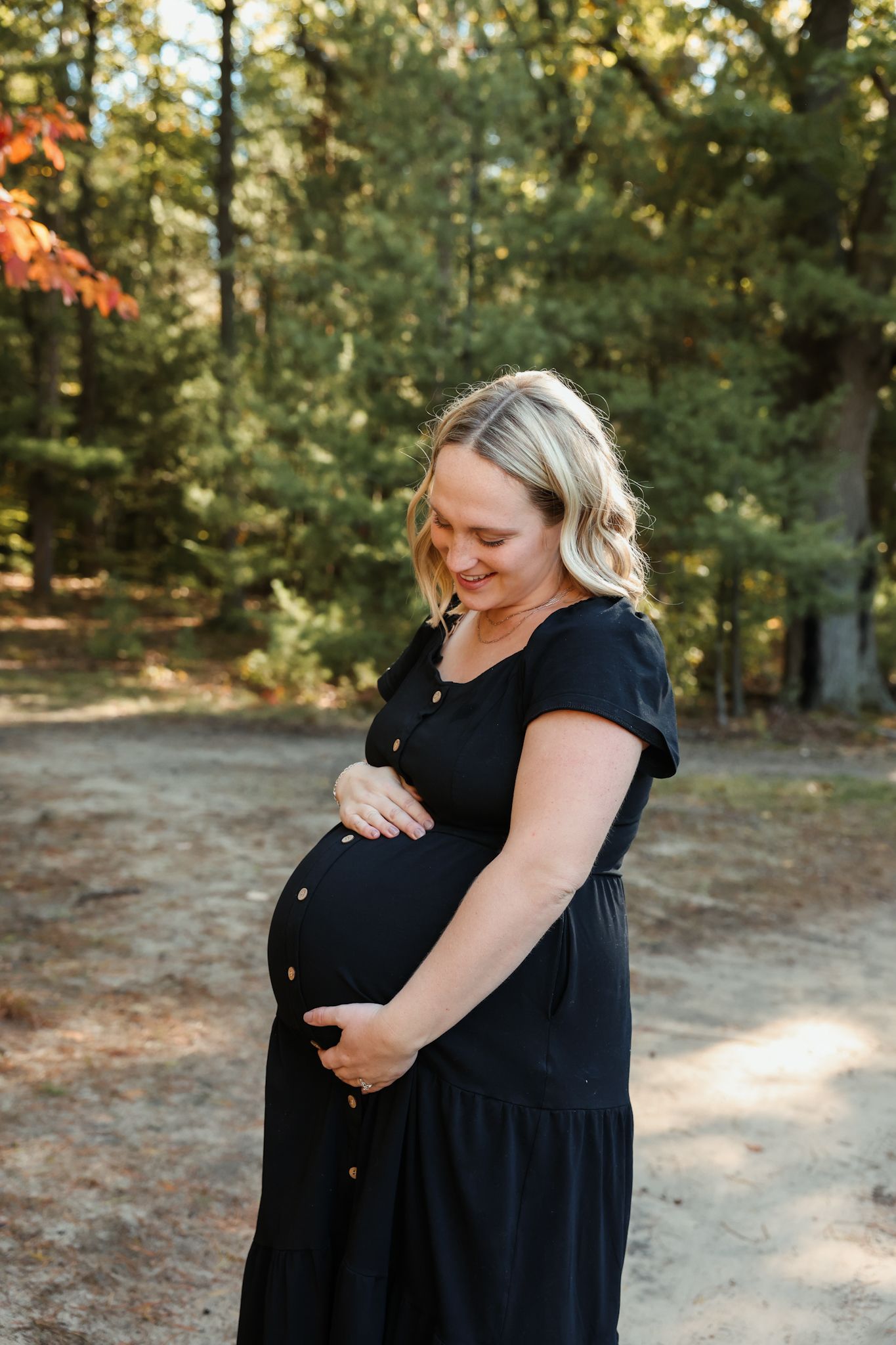 Smiling pregnant woman in black dress gently holding her baby bump outdoors in a sunny forest.